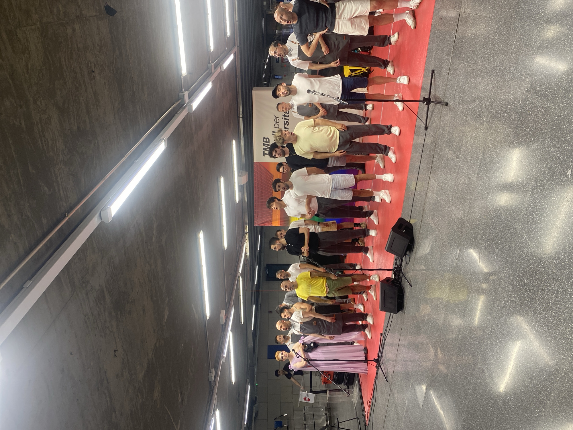 A group of approximately 15 men are standing on a raised red platform in what appears to be an indoor public space, possibly a metro station, in Barcelona, Spain. The men are dressed in various casual outfits, including t-shirts, shorts, and pants, with some wearing aprons. One person on the far left is wearing a long, light purple dress. The men are positioned in two rows, facing forward and some appear to be singing or performing. There are microphones and speakers set up on the platform, suggesting a performance. In the background, there are large advertisements or signage, including one with text that is partially visible and appears to say "TMB per...niversita". The ceiling of the space is concrete with industrial lighting fixtures. The floor is a polished grey surface.
