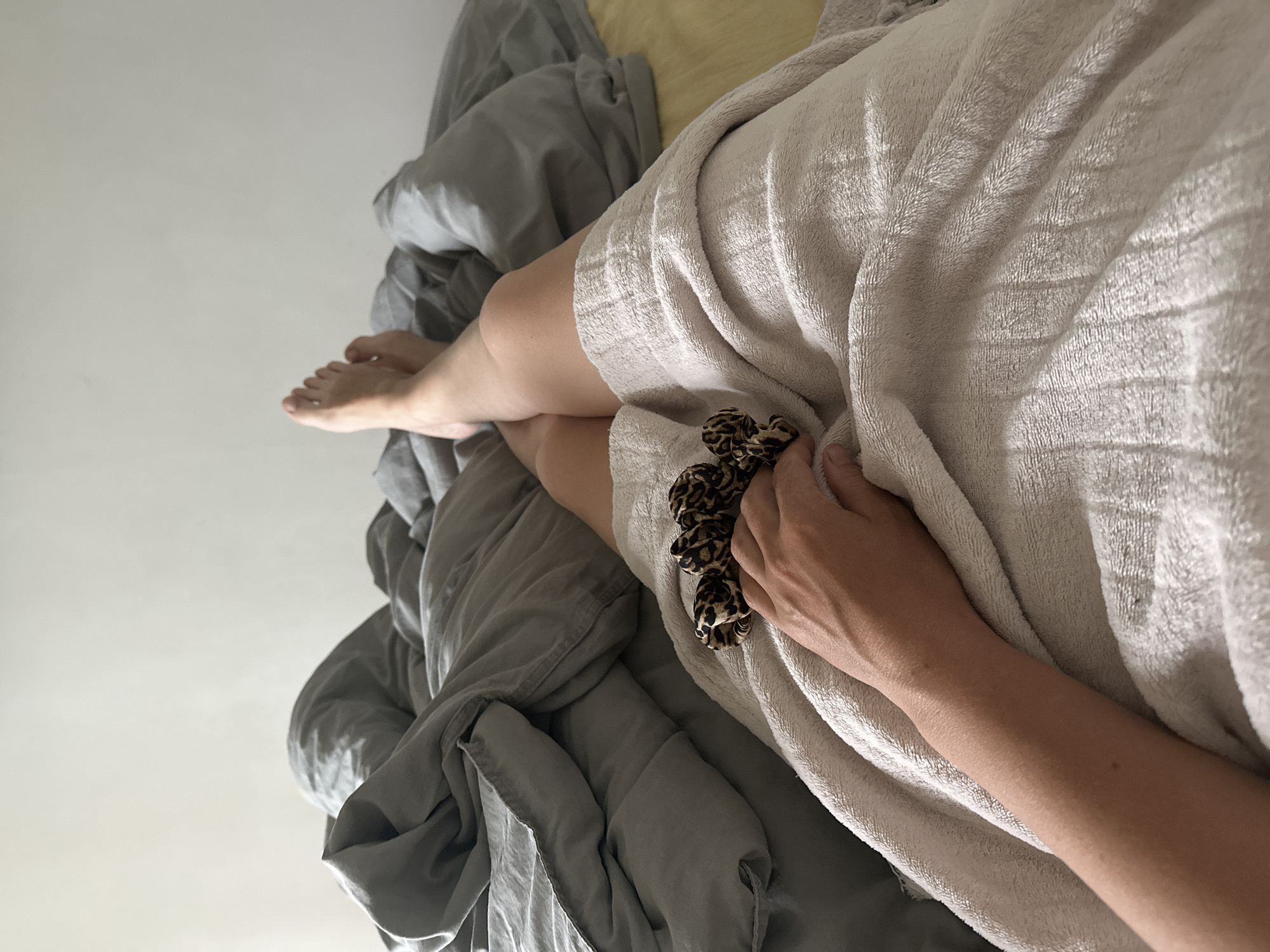 A person lies in bed with their legs extended, partially covered by a white bathrobe or towel. A hand holds a scrunchie with a leopard print pattern. The bedding consists of gray sheets and a yellow pillow. The scene is likely indoors. The city of Chaikovsky, Russia is not visually evident in the image.
