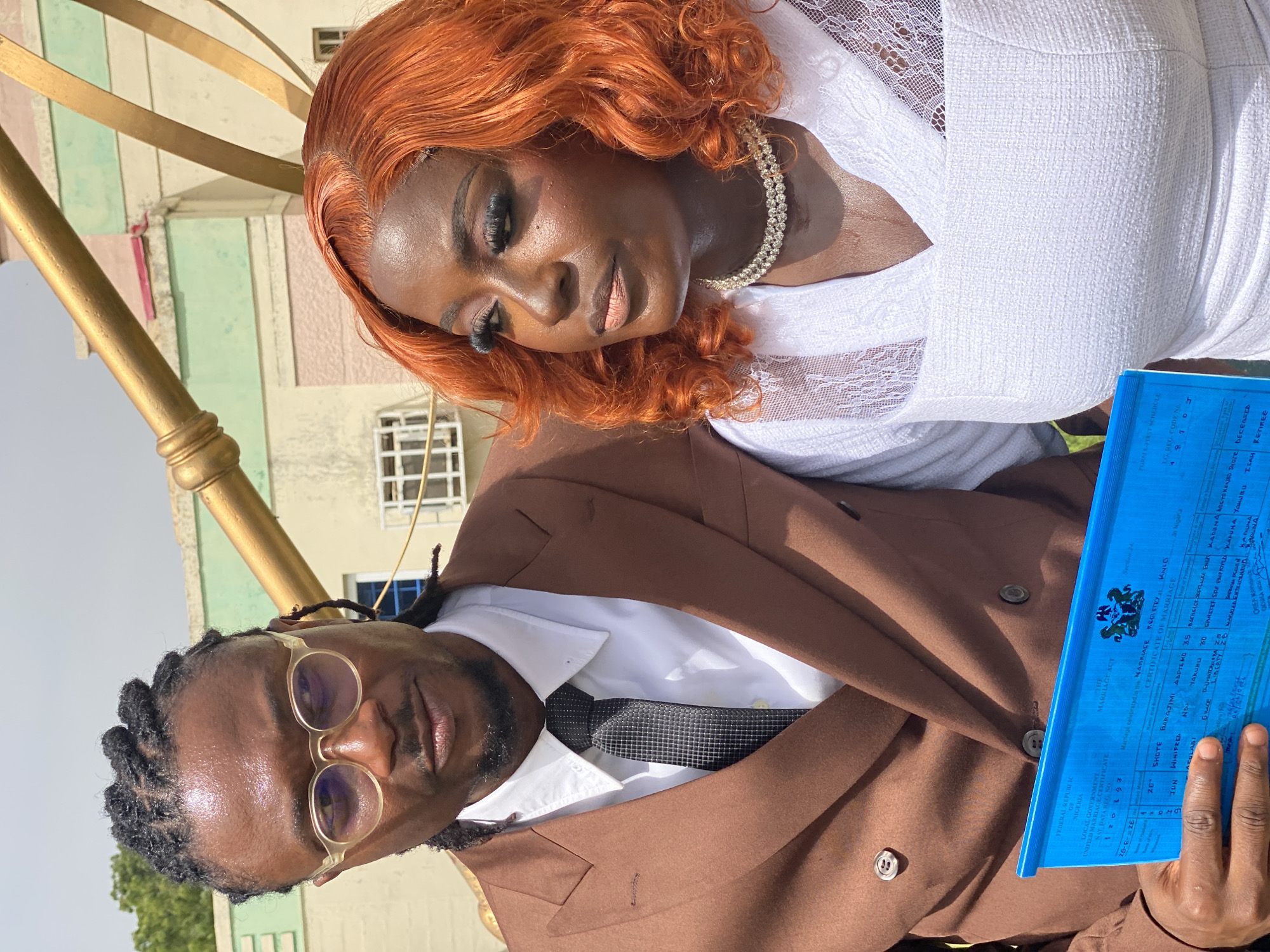 The image depicts a close-up, tilted shot of a couple, likely in Dadi Riba, Nigeria. The woman, in the foreground, has vibrant orange hair and is wearing a white lace-trimmed dress and a sparkling necklace. She has dramatic eye makeup and appears to be looking down, with a serene or thoughtful expression. The man is behind her and to the left, wearing a brown suit, white shirt, and a patterned tie. He has glasses with translucent frames and his hair is styled in dreadlocks. He is looking directly at the camera with a neutral expression. In his hands, he holds a bright blue document, which appears to be a marriage certificate. The text on the document is partially visible, indicating it's a "UNIFIED MARRIAGE CERTIFICATE" from the "FEDERAL REPUBLIC OF NIGERIA." The background shows a building with pastel-colored sections and windows, suggesting an outdoor setting with some architectural elements. The lighting indicates it is daytime.