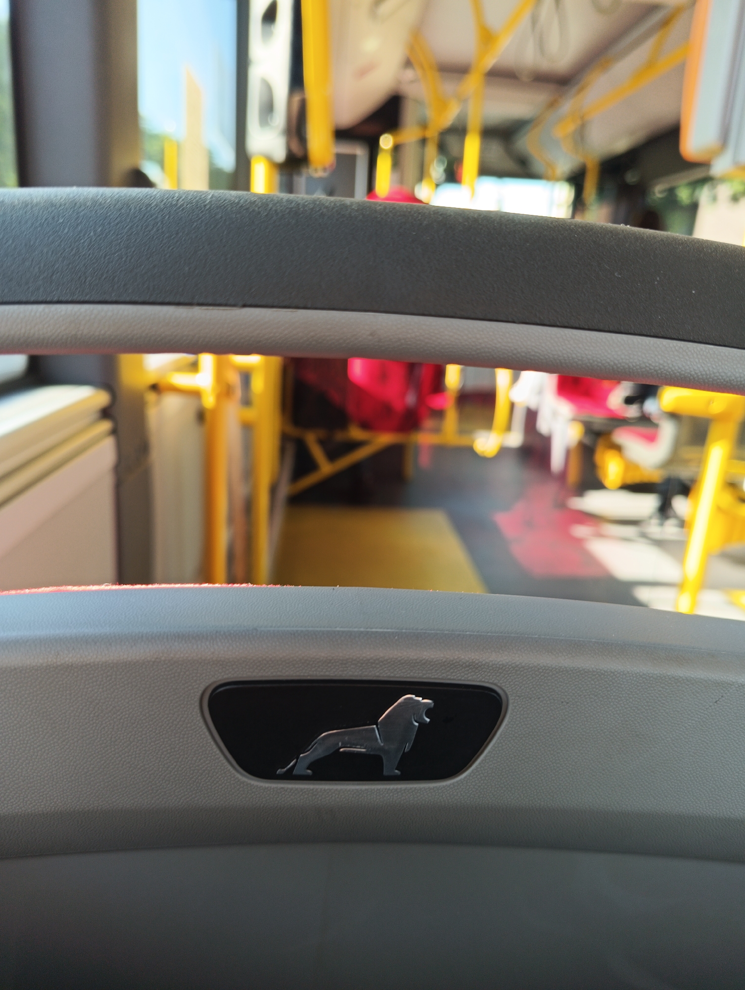 The image shows the interior of a bus in Gdańsk, Poland. In the foreground, a textured grey panel features a black, oval-shaped emblem of a lion in silhouette. The lion is depicted in profile, with its mouth open and tail raised. The background is blurred, revealing the yellow poles and handrails typical of a public transport vehicle, along with red seating and windows. The overall impression is of the driver's or passenger's seat area, looking forward into the bus.