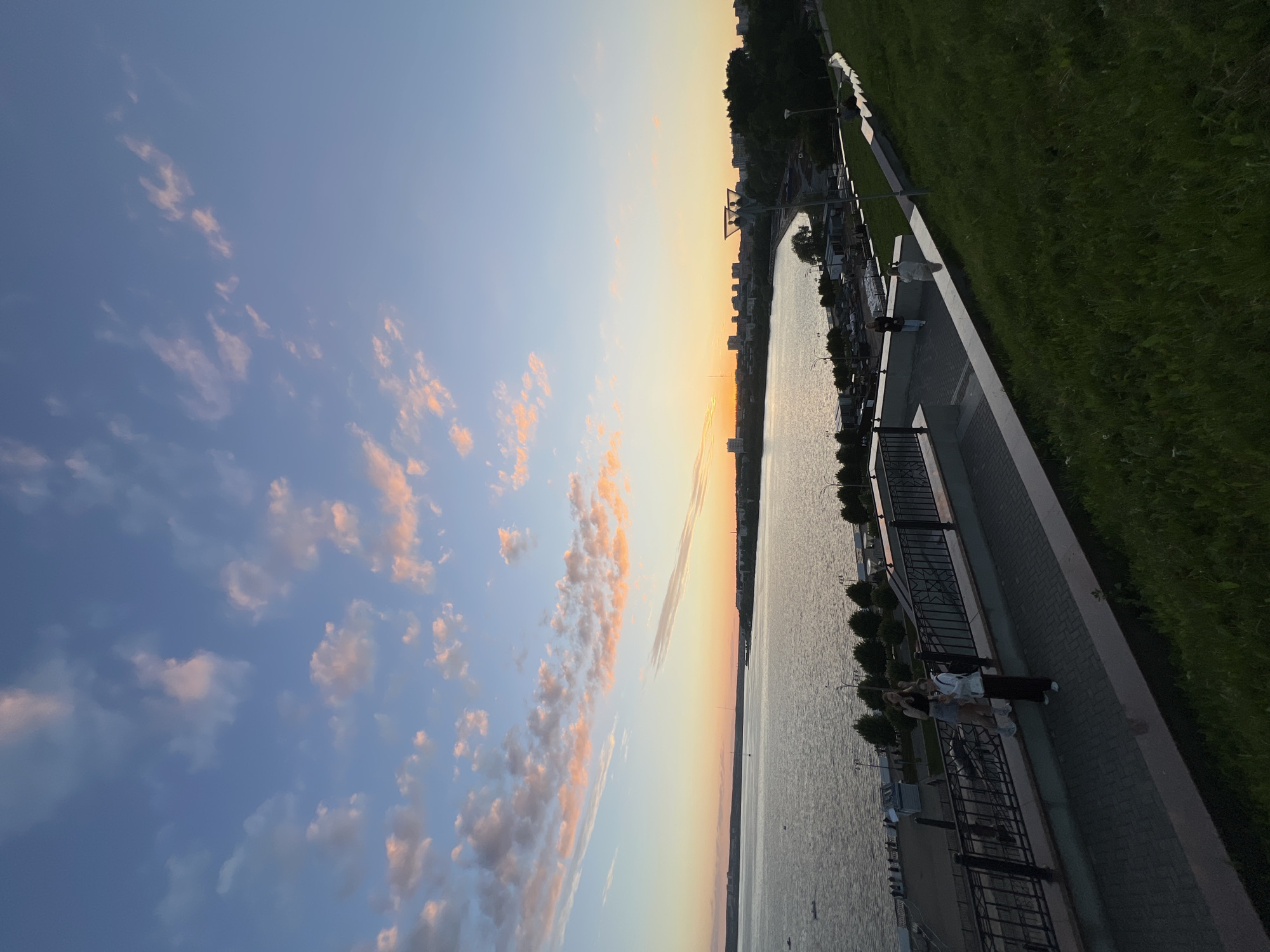 The image shows a scenic view of the Kama River in Izhevsk, Russia, at sunset. The sky is a vibrant gradient of blue, pink, and orange, with scattered clouds. The river is wide and calm, reflecting the colors of the sky. In the distance, the city skyline of Izhevsk is visible, with buildings and trees lining the shore. A paved walkway runs along the riverbank, with a black metal railing. Two young women are standing on the walkway, one taking a photo with her phone, and the other looking out at the river. The walkway is surrounded by a grassy slope, which is illuminated by the fading sunlight. There are streetlights visible along the walkway, and a few boats are dotted on the river. The overall atmosphere is peaceful and serene.