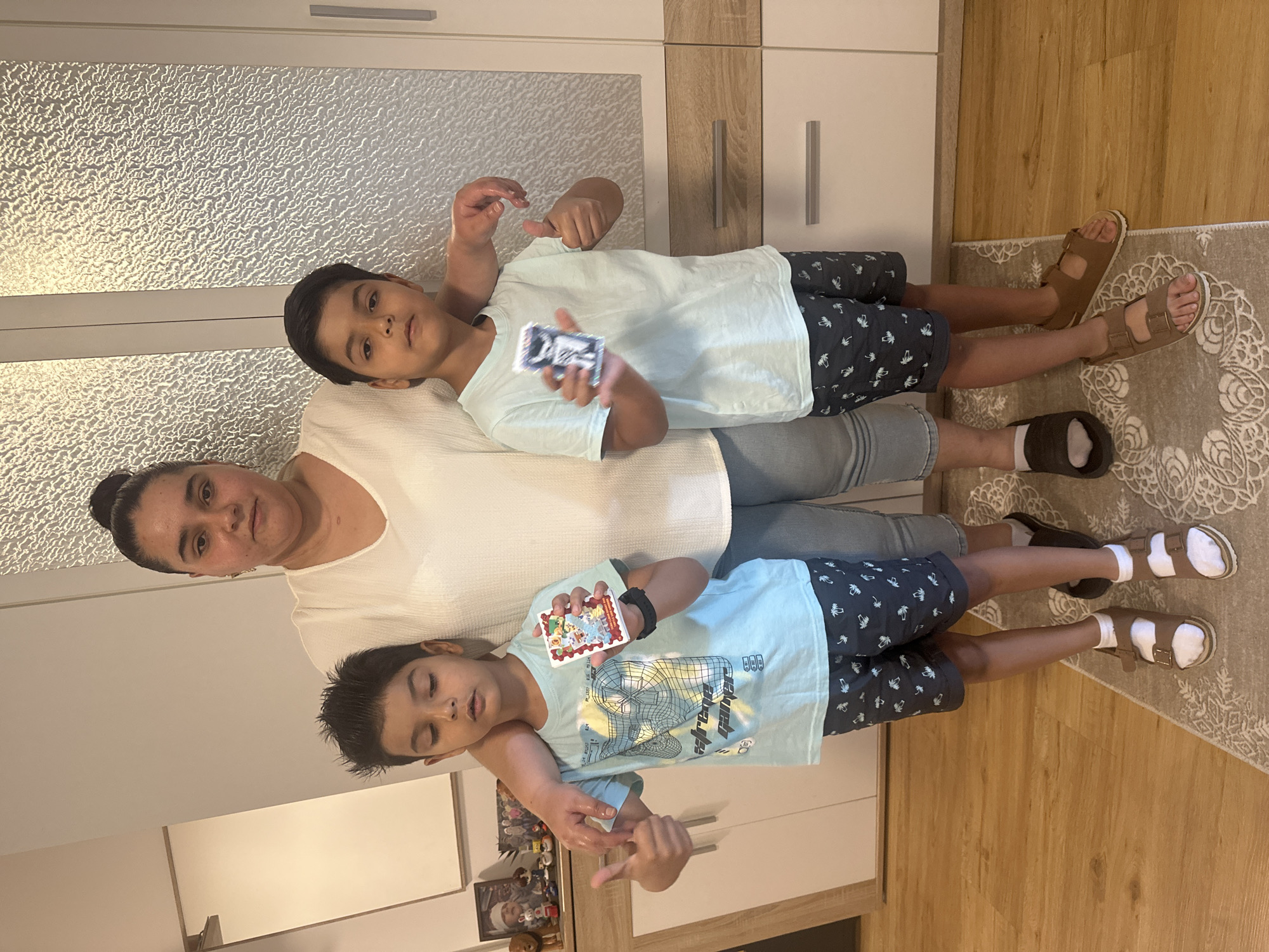 This image features a woman and two young boys indoors, likely in a home environment in Mainburg, Germany. The scene appears casual and domestic. The woman, with her hair pulled back, wears a white top and is positioned in the foreground, looking towards the camera with a neutral expression. She is holding one of the boys.
The two boys are positioned in front of and slightly behind the woman, with one boy leaning on her shoulder. The boy leaning on her shoulder is wearing a light blue t-shirt with a graphic print and dark shorts with a palm tree pattern. He is holding a small, rectangular object, possibly a card game or trading cards, with a colorful design. The second boy is standing in front of the woman and has his eyes closed. He is wearing a similar light blue t-shirt with a graphic design and dark shorts. He is also holding a small card. Both boys are wearing sandals with socks.
The background suggests a room with kitchen or closet cabinets, one of which has a textured glass door. The floor is covered with a patterned rug and wooden flooring. The lighting suggests it is daytime, but no specific cues about weather are apparent. The overall mood seems relaxed and possibly a moment captured during play or a shared activity. No visible text is clear enough to be transcribed.