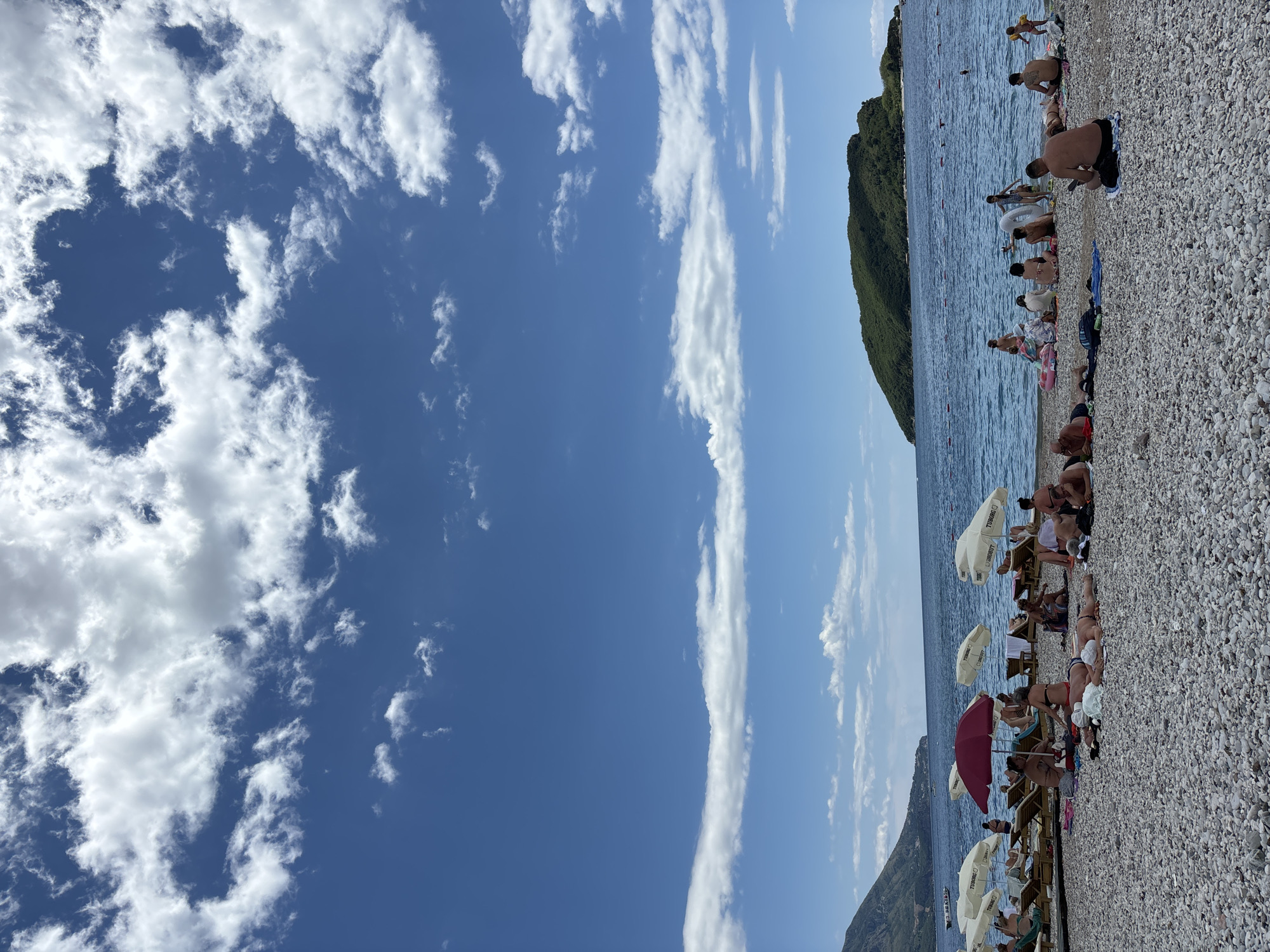 This is a wide-angle, full-frame, eye-level shot of a pebble beach in Budva, Montenegro. The sky is a vibrant blue with scattered, fluffy white clouds, some of which form a horizontal band across the mid-horizon. The sea is calm and blue, with a small, green, tree-covered island visible in the distance. The beach is populated with many people sunbathing, sitting, and relaxing on lounge chairs and towels. Several white umbrellas are set up for shade, along with one red umbrella. The people are mostly dressed in swimwear, and their activities include sunbathing, conversing, and looking at their phones. The pebble texture of the beach is prominent in the foreground. The overall impression is a lively and sunny beach scene on a summer day.