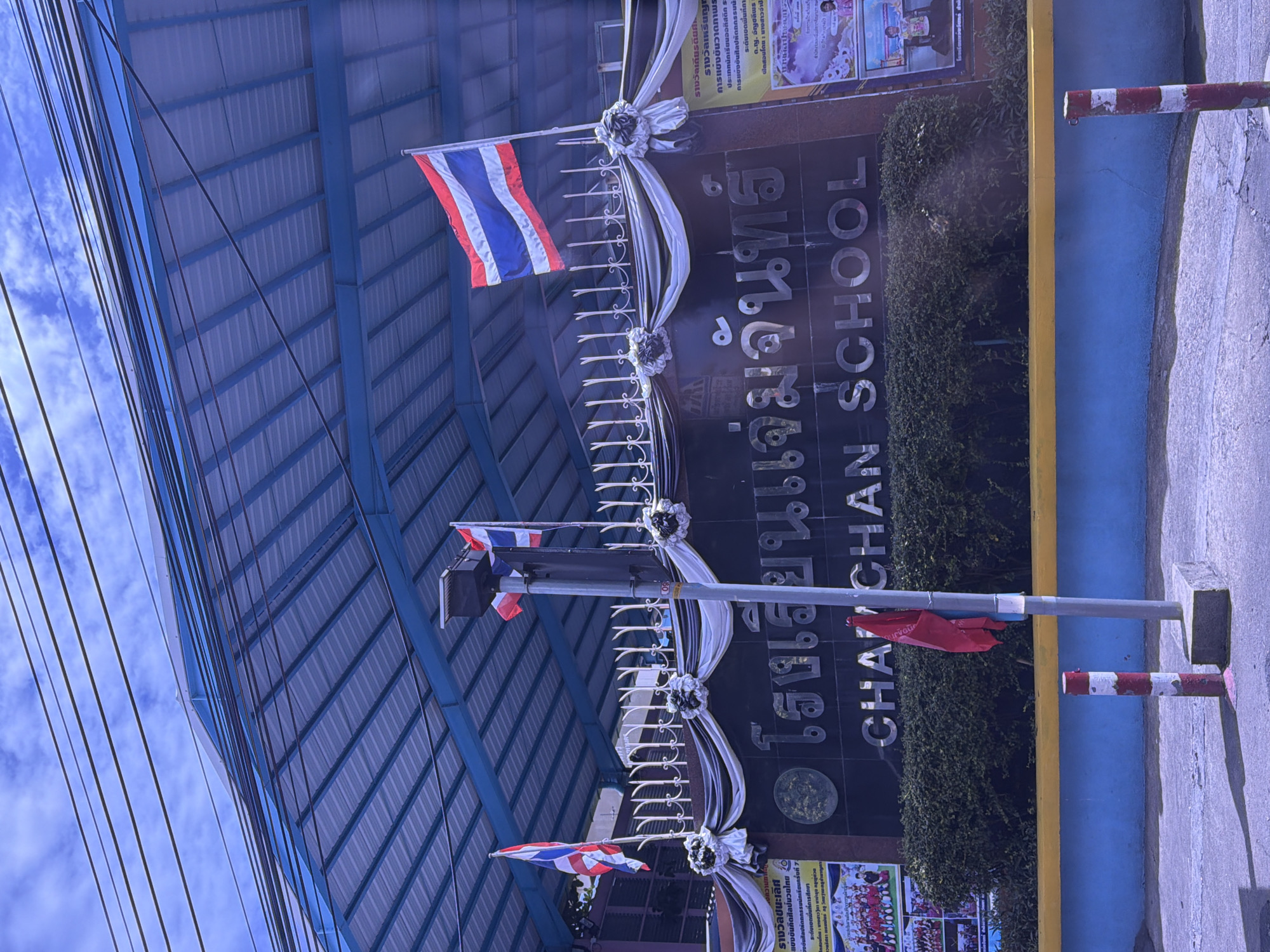 The image shows the exterior of "CHAI CHEN SCHOOL" in Bangkok, Thailand. The school building has a large, dark-colored facade with the school name prominently displayed in silver lettering. Above the school name, Thai lettering is also visible. The building is covered by a blue, corrugated metal roof supported by a metal frame. Three flags are visible: two Thai flags flying from poles on the roof, and a red flag attached to a lamppost in front of the school. The lamppost is gray with a solar panel on top. In front of the school is a manicured hedge. A yellow and blue painted wall runs along the bottom of the image, with a gray concrete sidewalk in front of it. Two red and white striped bollards are present on the sidewalk. The sky above is blue with scattered white clouds, and overhead power lines are visible. There are posters on the right side of the building, with text and images that are partially visible. One poster appears to depict students in uniforms. The image has a bright, sunny feel, though the lighting suggests it may be mid-day.