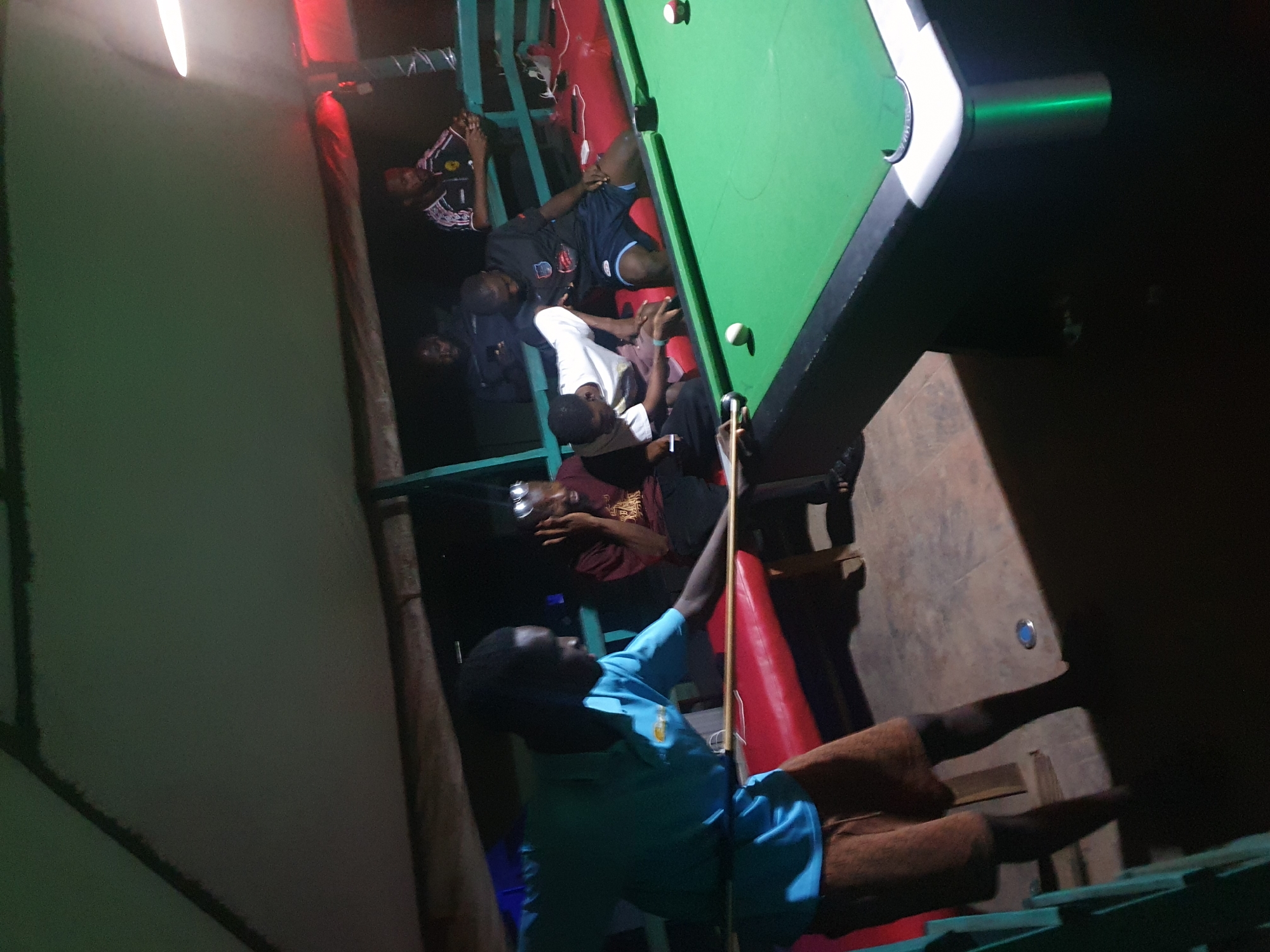 In Lokoja, Nigeria, a group of young men are gathered around a pool table in an outdoor, dimly lit recreational area, possibly an open-air bar or lounge. The scene is illuminated by a mixture of overhead lighting, casting shadows and highlighting the green felt of the pool table.
The focal point is a man in a turquoise polo shirt and brown shorts, actively playing pool. He is positioned to the left, leaning over the table with a pool cue extended, preparing to strike a white ball. His gaze is concentrated on the game.
Other individuals are dispersed around the table, observing the game or engaged in conversation. To the right of the player, a man in a maroon t-shirt and black shorts is seated, with his hand to his face as if in thought or amusement. Further back, several other men are visible. One, in a black t-shirt with a red emblem and blue shorts, is seated, his attention directed towards the pool table. Another man, wearing a black and white patterned sports jersey, is also seated, looking towards the game. Two other individuals are partially visible in the background, one appearing to be looking at his phone.
The environment suggests a casual, social gathering. A red bench runs along the side of the pool table, and the flooring appears to be concrete or tiled. The overhead structure, possibly a thatched roof or canopy, is visible, contributing to the outdoor setting. The overall atmosphere is one of relaxed camaraderie, centered around the game of pool.