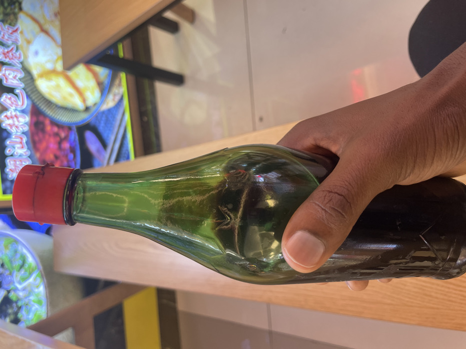 A person's hand is holding a glass bottle of dark liquid with a red cap. The bottle is made of dark green glass and is partially obscured by the hand. The bottle is being held in a restaurant. The background shows a portion of the restaurant’s decor. There are wooden tables and chairs in the background. On the wall behind the bottle, there is a poster with Chinese characters. The poster has a picture of a dish on a plate, which is partially visible. This location is a restaurant located at coordinates 29.566, 106.582 in Chongqing, China.