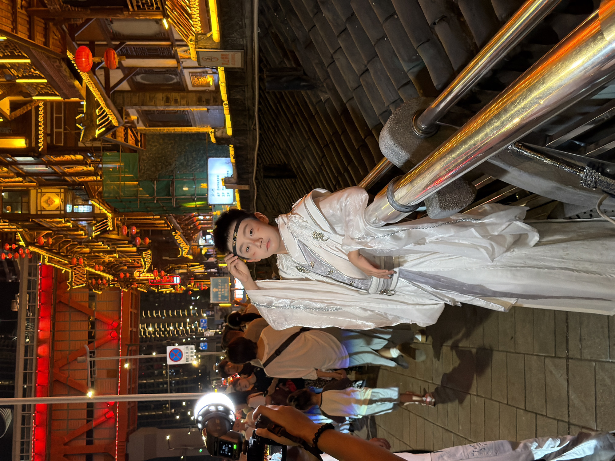 An individual, appearing to be a child, stands on a paved walkway at night, wearing a white, flowing garment with silver trim, possibly a traditional costume. The child has a black headband and makeup on their face. They are leaning on a metal railing that overlooks a street lined with traditional buildings illuminated with yellow and red lights. In the background, a large red illuminated bridge structure is visible. Other people are walking on the walkway and in the background. The scene appears to be in a busy urban area at night. The location is identified as Yuzhong District, China.