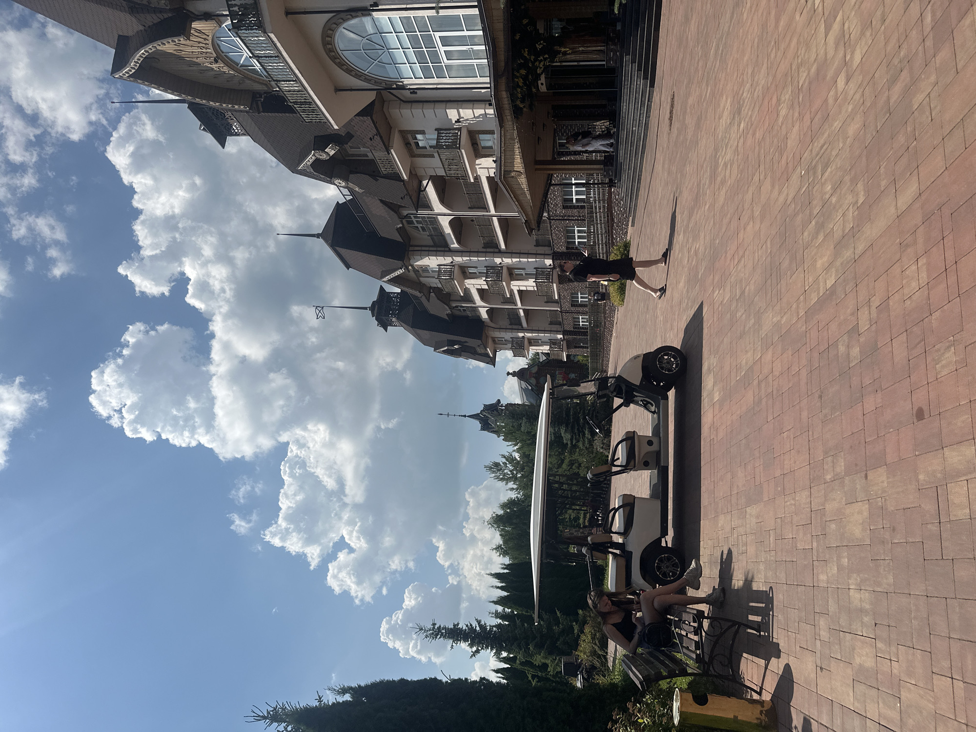 In Bogdanikha, Russia, a resort complex is captured on a sunny day with a bright blue sky filled with cumulus clouds. In the foreground, a beige golf cart is parked on a red brick pathway. To the left of the golf cart, a young woman with blonde hair wearing a black tank top and shorts sits on a park bench, looking down at her phone. To the right of the golf cart, a person in a black dress and sandals walks away from the camera towards a large, ornate building with multiple balconies, arched windows, and dark, sloping roofs. The building appears to be a hotel or resort. In the background, numerous tall evergreen trees surround the area, and a dark, spire-like structure is visible behind the main building. The overall scene depicts a serene and luxurious outdoor environment.