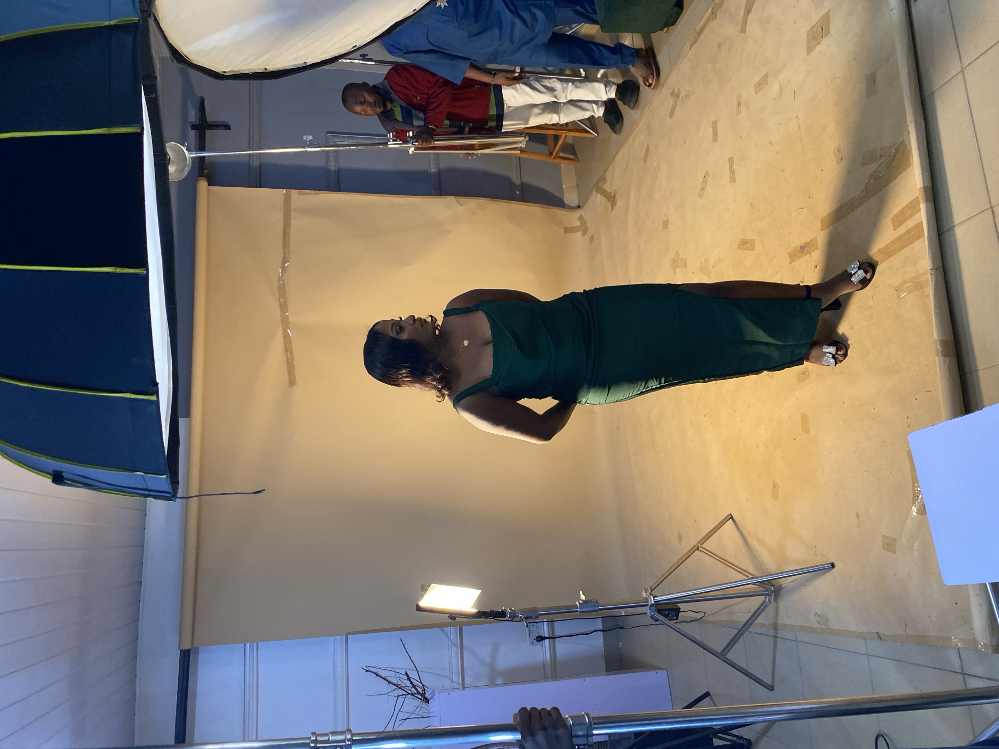 In Bukuru, Nigeria, a woman in a form-fitting, dark green, floor-length dress with a thigh-high slit stands posing in a studio setting. She wears black open-toed heels with embellished straps. Her hands are clasped behind her back, and she looks to her right, away from the camera. The studio has a light beige backdrop that appears to be paper or fabric, which is taped to the floor in several places. Several studio lighting equipment pieces are visible, including a large softbox light and a smaller LED panel light on a stand. Two men are present in the background; one stands to the right of the woman, wearing a red jacket with green and blue stripes, white pants, and black shoes. Another man, partially obscured, stands further to the right in a blue garment. A metal light stand is in the foreground to the left, and a white board is on the floor in the bottom right corner. The overall lighting is bright, characteristic of a professional photo shoot.