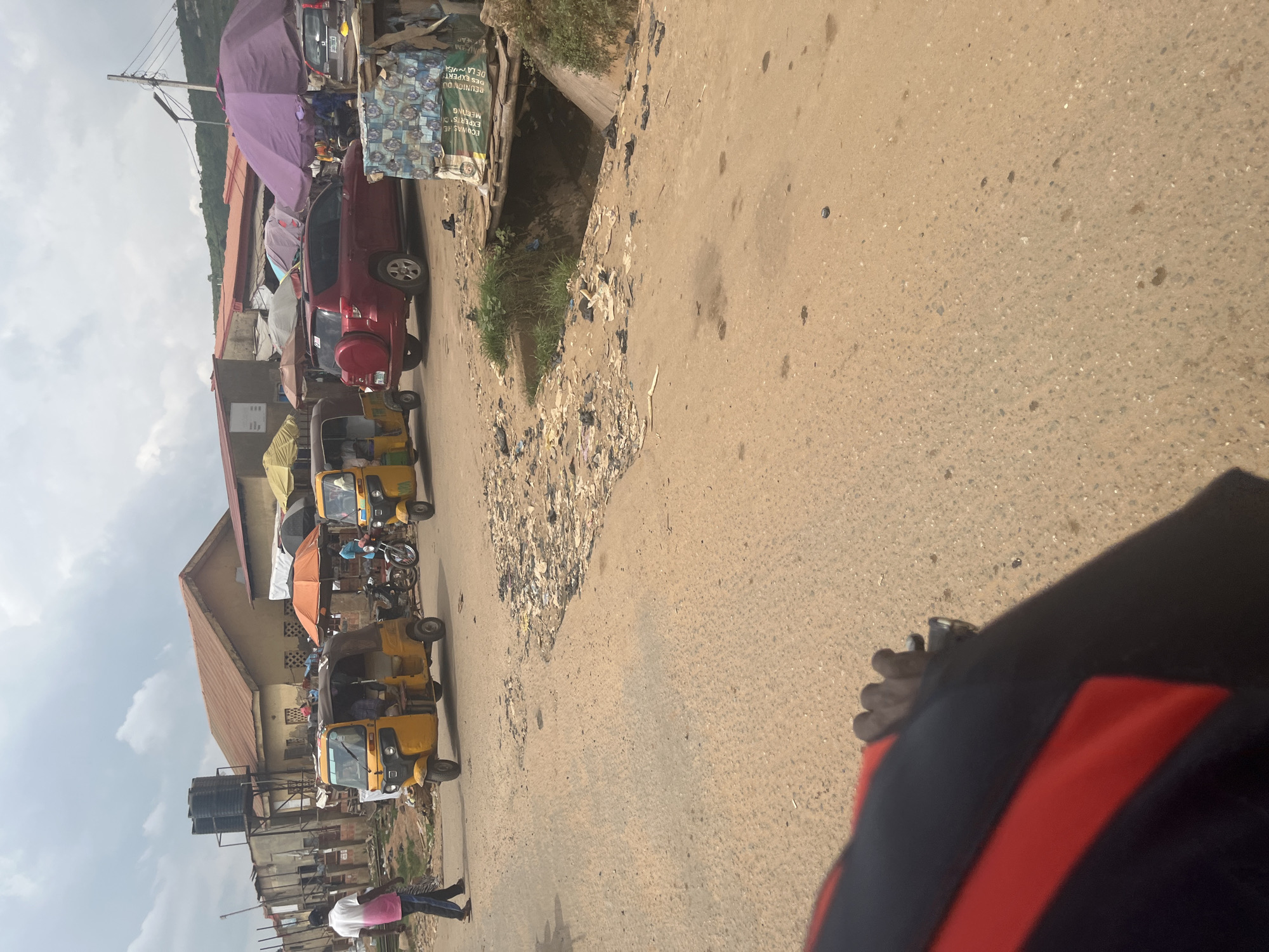 A medium shot captures a street scene in Abuja, Nigeria, on a cloudy day. The foreground is dominated by a portion of a person's arm, clad in a black and red jacket, holding onto handlebars that are not visible. The road surface is unpaved and appears dusty, with scattered debris and some puddles or stains.
In the mid-ground, a yellow auto-rickshaw, known locally as a 'Keke Napep', is parked on the right side of the road, facing left. Another similar auto-rickshaw is visible behind it, also yellow. To the left, a person wearing a white shirt with a pink accent and dark trousers walks away from the camera. Behind the auto-rickshaws, a red SUV is parked. Several market stalls are visible in the background, with various colorful umbrellas providing shade. These stalls are constructed from corrugated metal and wood. A large purple umbrella is prominently displayed over one of the stalls. In the far background, a hillside covered in vegetation rises under the overcast sky. A large water tank on a metal structure is situated to the left of the buildings. The overall environment suggests a bustling, informal market area.