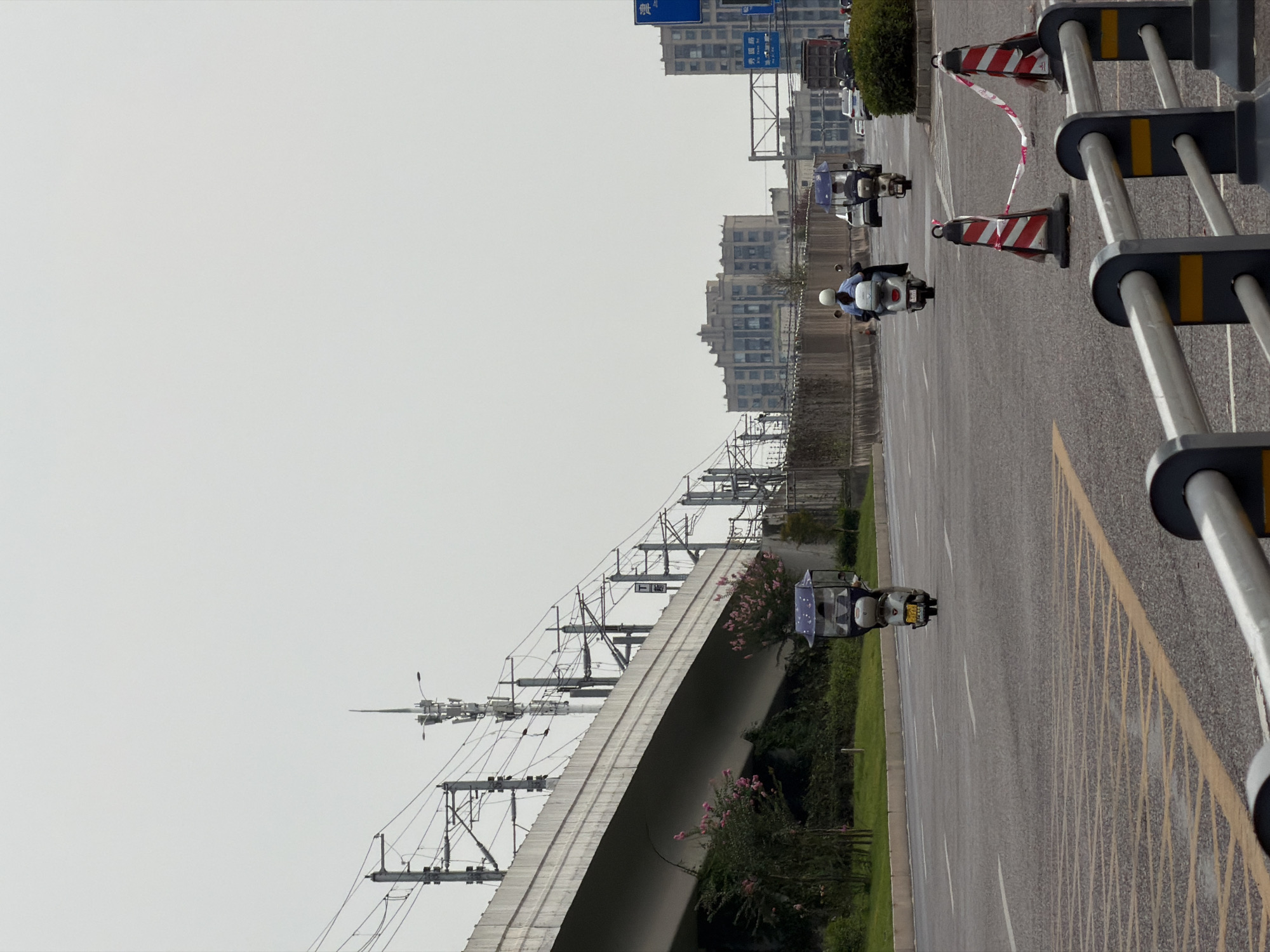 The image shows a street scene in Jiangjin, China. In the foreground, there are several traffic cones with red and white striped tape strung between them, indicating a temporary closure or warning. A silver railing with black and orange accents runs horizontally across the lower portion of the frame. The road surface is asphalt with white lane markings and yellow cross-hatching in some areas.
In the middle ground, three-wheeled vehicles and scooters are traveling on the road. The first vehicle from the left is a blue three-wheeled scooter with a canopy and a driver. To its right, a scooter with a white helmet is being ridden by a person. Further to the right, another three-wheeled vehicle with a canopy, similar to the first one, is also visible.
The background features a raised overpass with extensive electrical wiring and support structures, likely for a train or monorail. Beyond the overpass, there are several multi-story buildings, some of which appear to be apartment complexes or office buildings. The sky is overcast, casting a diffused light over the scene. Lush green vegetation is present along the side of the road beneath the overpass. A traffic sign with Chinese characters is visible on a pole to the right side of the road.