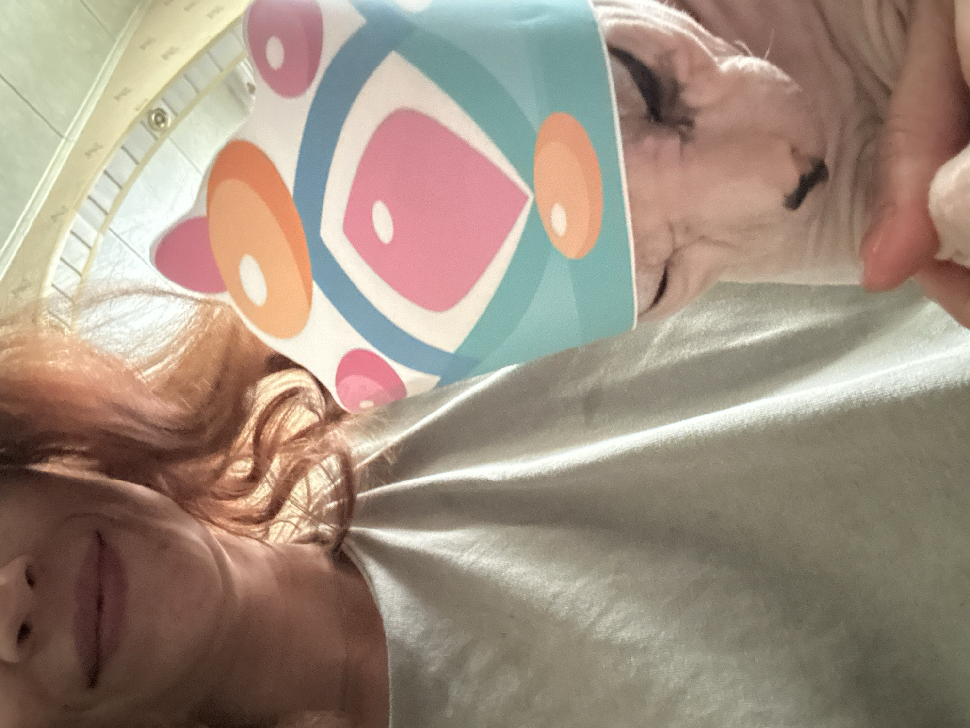 The image shows a close-up of a woman and a Sphynx cat. The woman, with red hair, is smiling and wearing a light gray t-shirt. The cat is wearing a colorful party hat. The background appears to be indoors, possibly a home, with a tiled ceiling visible. No specific landmarks of Togliatti, Russia are present in the image.