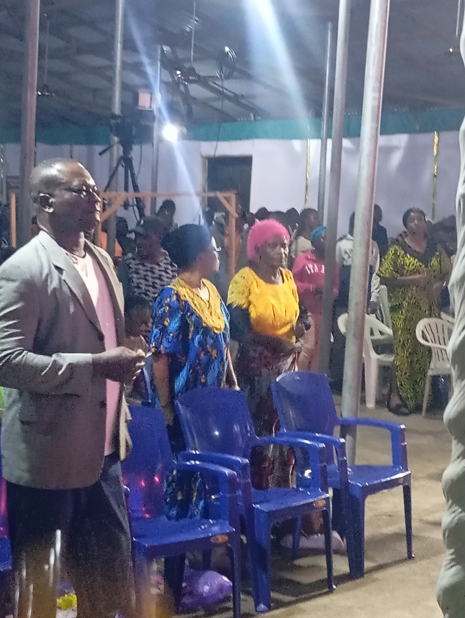 An outdoor gathering is taking place in Markurdi, Nigeria, under a partially covered structure. In the foreground, a man in a gray suit jacket and pink shirt stands on the left, looking to his right. Next to him, a woman in a blue and yellow patterned dress is also facing right. Further back, a woman with bright pink hair and a yellow top stands in the center. To her right, a woman in a green patterned dress is visible. Blue plastic chairs are arranged in rows, some empty and some occupied by attendees. The environment appears to be a community event or service, with a camera on a tripod visible in the background.