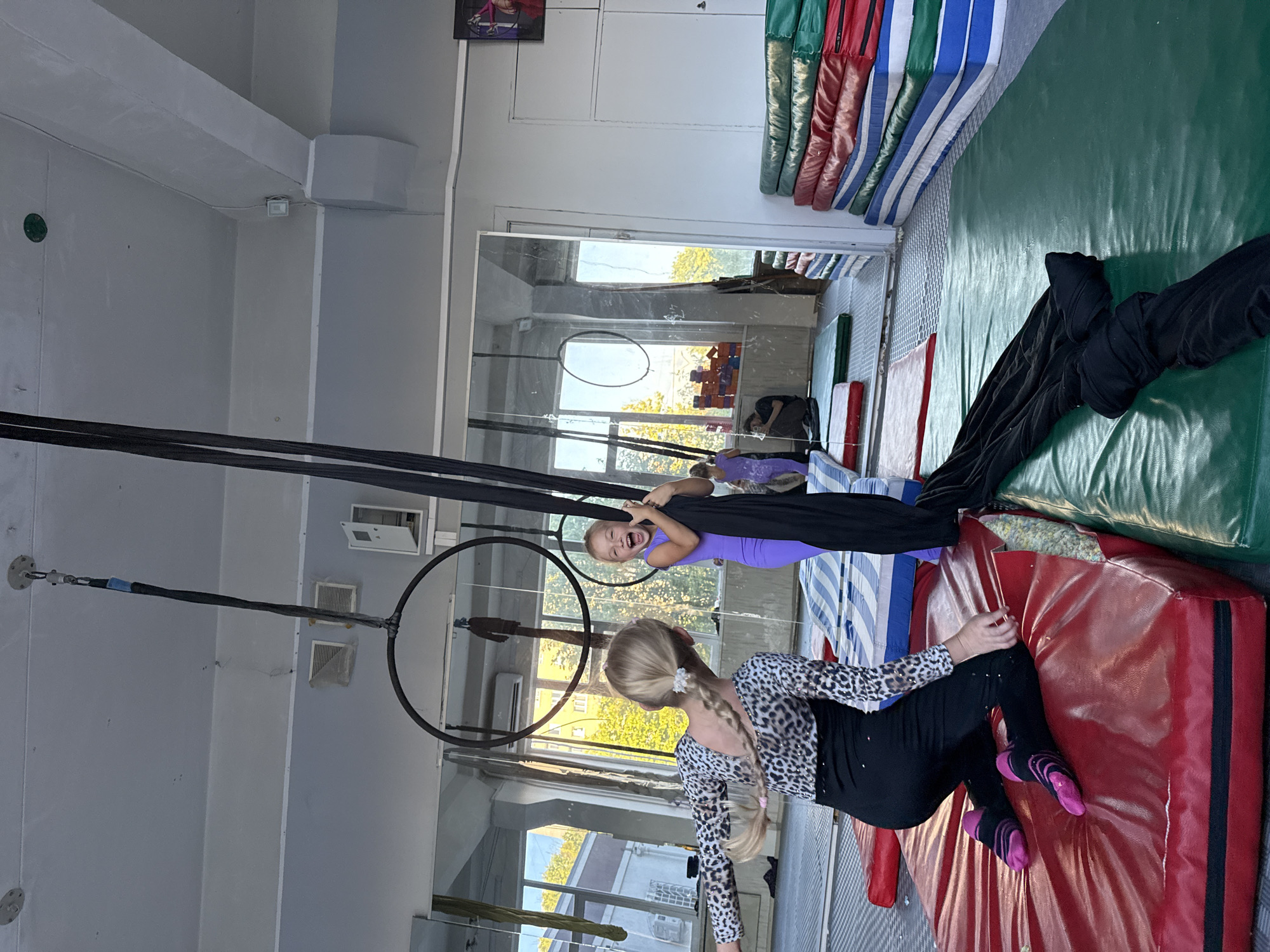 In a brightly lit indoor space, possibly a gymnasium or dance studio, two young girls are engaged in aerial acrobatics. The setting features high ceilings, large windows that allow natural light to filter in, and what appears to be a mirrored wall reflecting the scene. The floor is covered with padded mats in red, green, and blue, suggesting a safe environment for athletic activities. Several gymnastic apparatus are visible, including large hoops and black aerial silks.
One girl, with blonde hair styled in a braid adorned with a white flower, is dressed in a leopard-print top and black leggings. She is leaning on a red mat, her body positioned as if mid-performance or practicing a pose. Her facial expression is not clearly visible.
The other girl, who appears younger, is suspended mid-air by black aerial silks. She is wearing a vibrant purple leotard and has an excited, open-mouthed expression, suggesting joy and enthusiasm. Her blonde hair is loose, and she is gripping the silks with her hands.
The overall atmosphere seems energetic and playful, characteristic of children practicing a sport or performance art. The presence of gymnastic equipment and the girls' attire indicate a training or practice session. The time of day appears to be daytime, as suggested by the natural light coming through the windows. The location is identified as Dnipro, Ukraine. No visible text is present in the image.