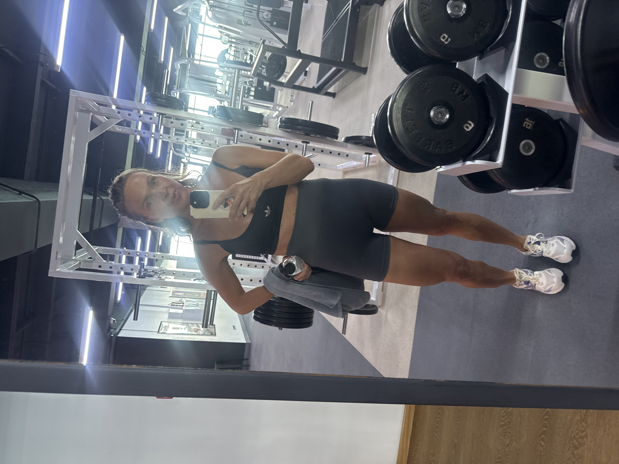 A woman in athletic wear takes a mirror selfie in a gym. She is wearing a dark sports bra and dark shorts, with white and blue sneakers. She is holding a water bottle and a gray towel in her left hand. In the background, there are weight racks, dumbbells, and other gym equipment. The lighting in the gym appears to be fluorescent and natural light from windows. The image was taken indoors at a gym.