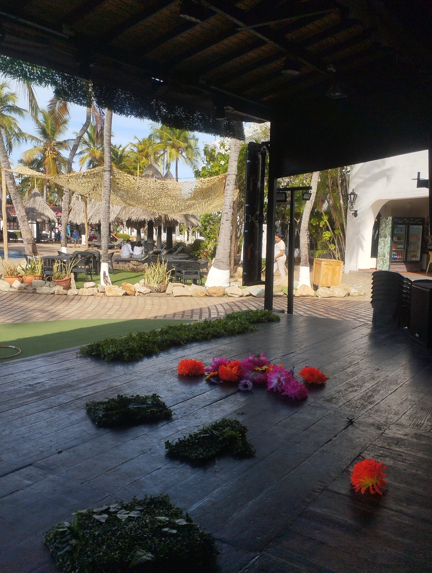 A partially shaded outdoor area features a dark wooden deck decorated with clumps of greenery and scattered orange and pink flowers. Beyond the deck, a patio area with palm trees, sunshades, and tables with people seated can be seen. To the right of the deck, there is a white building with an arched entrance and a beverage cooler. The setting appears to be a resort or event space, possibly in the vicinity of Porlamar, Venezuela, based on the tropical environment. The lighting suggests it is daytime.