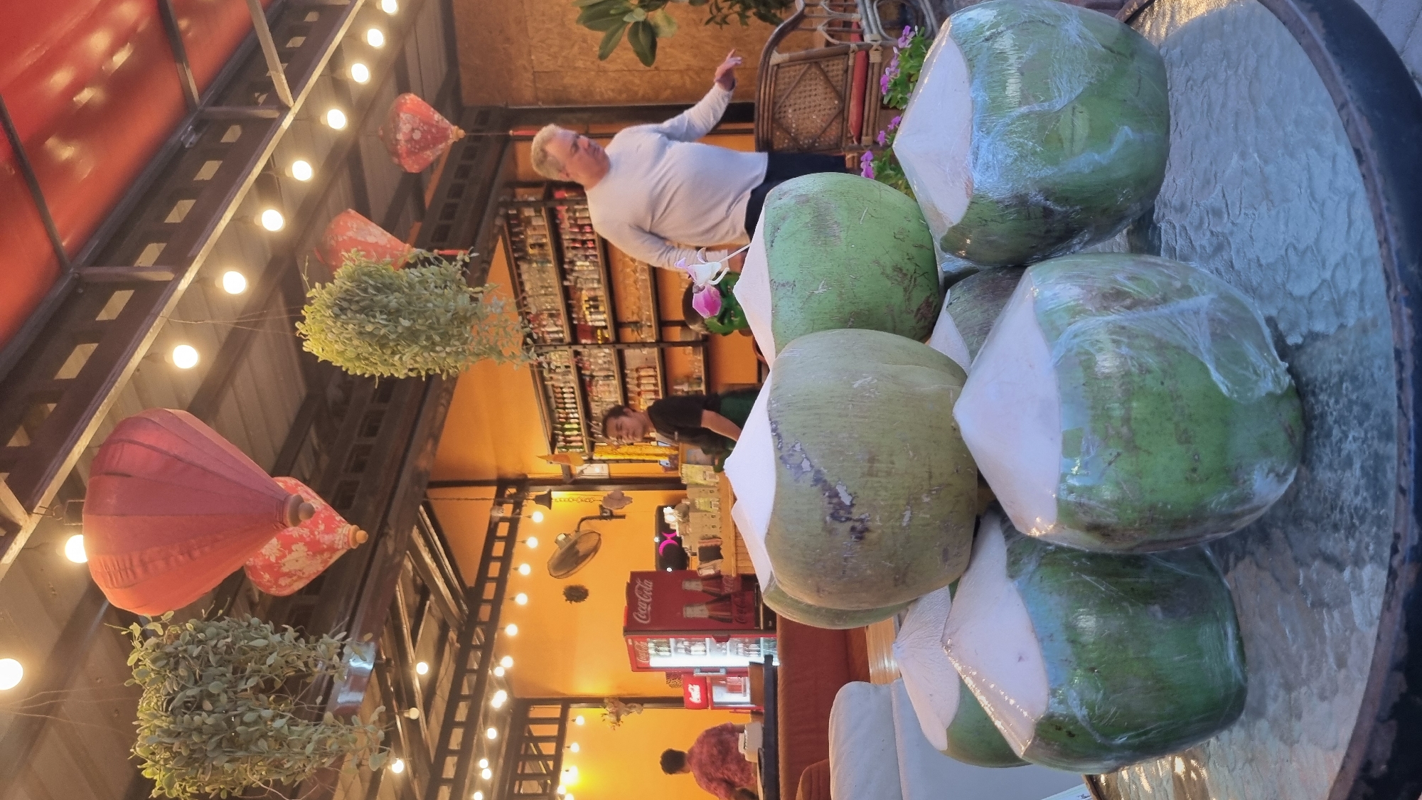 The image depicts a tropical setting, possibly a bar or restaurant in Pattaya City, Thailand. In the foreground, a collection of freshly prepared coconuts, their tops shaven and wrapped in plastic, are arranged on a glass-top table. The coconuts are a prominent subject, suggesting a refreshing beverage offering.

In the background, the establishment is visible with its warm orange-toned walls and a well-stocked bar featuring an array of bottles. A Coca-Cola refrigerator stands to the left. Two individuals are present: a man in a light-colored long-sleeved shirt is walking towards the right side of the frame, appearing to interact with the environment, while another person, likely staff, is standing behind the bar. Overhead, decorative red lanterns and hanging plants add to the ambiance, illuminated by string lights. The overall scene conveys a relaxed and welcoming atmosphere typical of a tropical vacation destination.