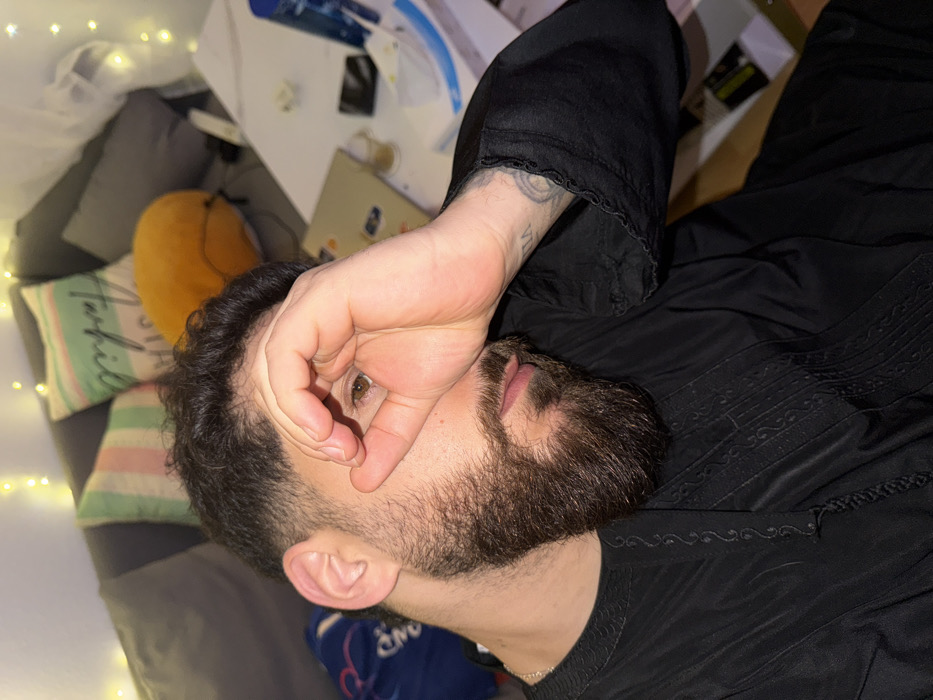 A man is depicted in a close-up, high-angle shot, lying down indoors, possibly on a couch or bed. He is positioned with his head towards the bottom of the frame and his body extending upwards to the right. His right hand is raised, partially covering his face, with his thumb and index finger forming a circle, allowing his right eye to peek through. His visible eye is brown. He has dark, curly hair, a full beard, and a mustache.
He is wearing a dark, likely black, long-sleeved garment that features intricate, geometric embroidered patterns on the chest and lower sleeve areas. A tattoo with a circular design and faint text or numbers is visible on his left forearm, just beneath the cuff of his sleeve.
The background suggests an indoor setting, likely a casual living space or bedroom. To the left of the man, several cushions are visible. One is striped in light green, pink, and white, with partially visible text that appears to spell out "Farfil" or similar. Another cushion is a solid orange or yellow color. Behind and above the man's head, a light-colored surface, possibly a table or desk, holds various items including papers and what looks like a mobile phone. String lights, often referred to as fairy lights, are draped in the top-left and bottom-left portions of the frame, emitting a warm, soft glow, suggesting the photo was taken during the evening or at night. The overall impression is one of a relaxed, intimate moment.