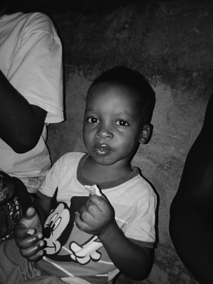 A young child wearing a shirt with a Mickey Mouse cartoon on it. The child is holding a small piece of food and looking directly at the camera. The background is plain and textured, with a portion of another person visible to the left and another to the right of the child. The image is in black and white.