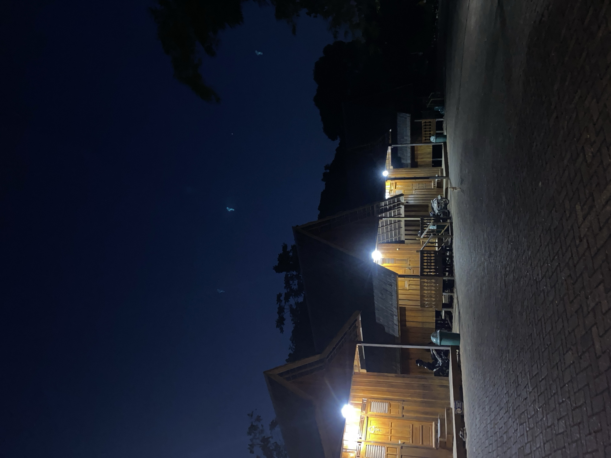 The image depicts a row of wooden cabins at night, illuminated by exterior lights. The cabins have steeply pitched roofs and wooden doors with decorative grilles. A paved walkway made of interlocking bricks runs in front of the cabins. A green trash bin is visible next to one of the cabins, and a motorcycle is parked near the entrance of another. In the background, silhouetted trees and a hillside are visible against a dark blue sky. There are faint, blurry light anomalies in the sky that could be lens flares or natural phenomena. A person is seated on a dark object to the left of the frame, near the steps of one of the cabins. The overall scene suggests a tranquil, possibly rural, setting in Indonesia.