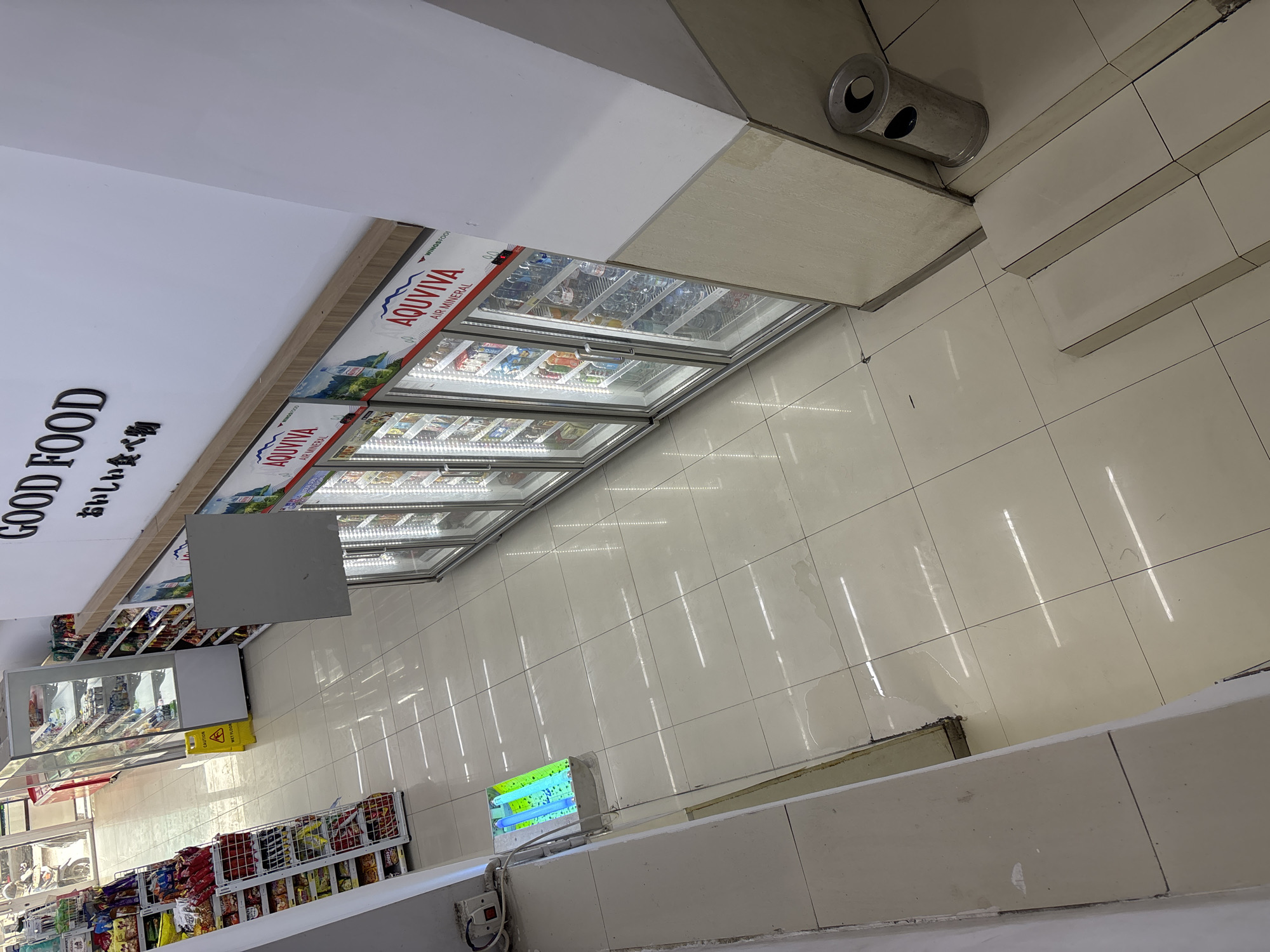 The media file presents an interior view of a grocery or convenience store, likely located in West Jakarta, Indonesia. The image is captured from an elevated perspective, looking down a wide aisle with a clean, light-tiled floor that reflects the overhead artificial lighting.
A prominent feature is a long row of chest freezers or refrigerators with glass tops, extending along one side of the aisle. These freezers contain various packaged food items and beverages, illuminated by internal lights. The branding "AQUVIVA AIR MINERAL" is clearly visible on the lids of these units, alongside smaller text that appears to be "WINGSFOOD". Above this row of freezers, a white wall displays the words "GOOD FOOD" in large black letters, accompanied by Japanese text "おいしい食べ物" (Oishii tabemono), meaning "delicious food".
Opposite the freezers, to the right of the aisle, several metal shelves are visible, stocked with a variety of snack bags and other packaged goods. A yellow "CAUTION WET FLOOR" sign stands near a smaller display unit in the mid-ground of the store. Further into the store, more product-filled shelves can be seen. Near the top right corner of the frame, a metallic cylindrical object, possibly an ash tray or trash bin, is mounted on a surface. Closer to the foreground on the right, a small light fixture with two fluorescent tubes is visible, positioned near a low white wall or counter. The store appears well-lit and organized. No people are present in the image.
