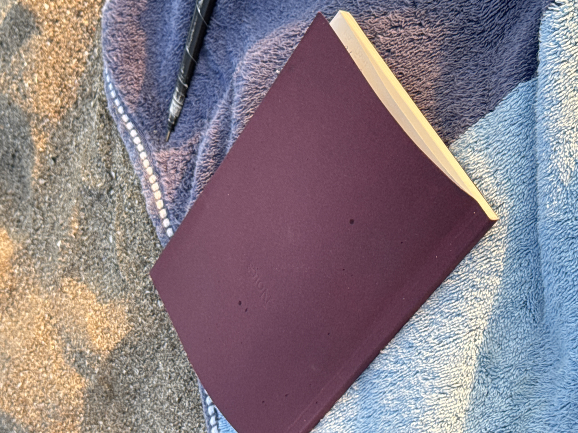 A closed, dark red notebook with the embossed word "Notes" on its cover lies on top of a blue towel. To the right of the notebook, a black and white marbled pen rests on the towel. The towel is partially on a sandy surface, illuminated by soft, warm light. The image is taken from a high angle, looking down. The location appears to be a beach in North Kuta, Indonesia, given the sand and towels indicative of a beach setting. There are no people visible in the frame.