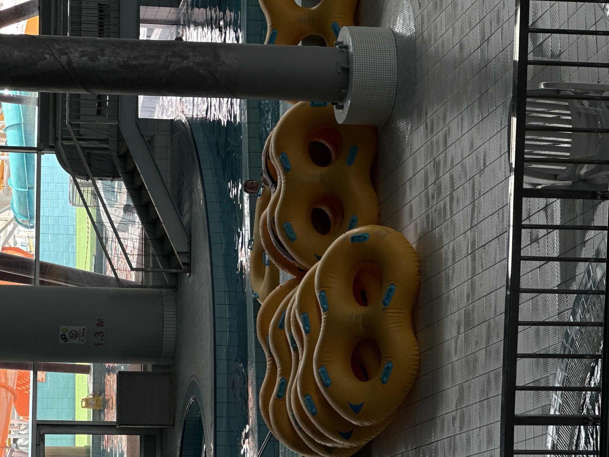 An indoor water park in Minsk, Belarus, features a swimming pool with inflatable tubes stacked on the tiled floor. A person is visible in the pool. In the background, a staircase leads to a waterslide. A pillar with a depth marker indicating "1.3 M" stands near the pool.