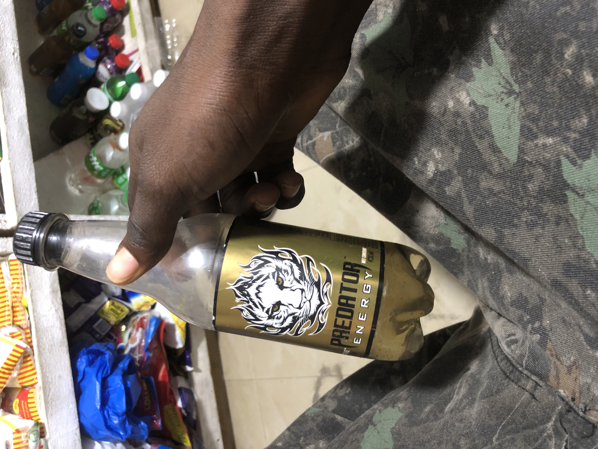 A close-up shot shows a person's hand holding a clear plastic bottle of Predator Energy drink. The bottle has a prominent gold label with a stylized white tiger head and the words "PREDATOR ENERGY" in black text. The drink inside appears to be a golden-brown color. The background is blurred, but it suggests shelves stocked with various packaged goods and other bottles, possibly in a store. The lower part of the image shows the person's legs, which are clad in camouflage pants. The scene appears to be indoors, likely a small shop or market stall in Ado, Nigeria.