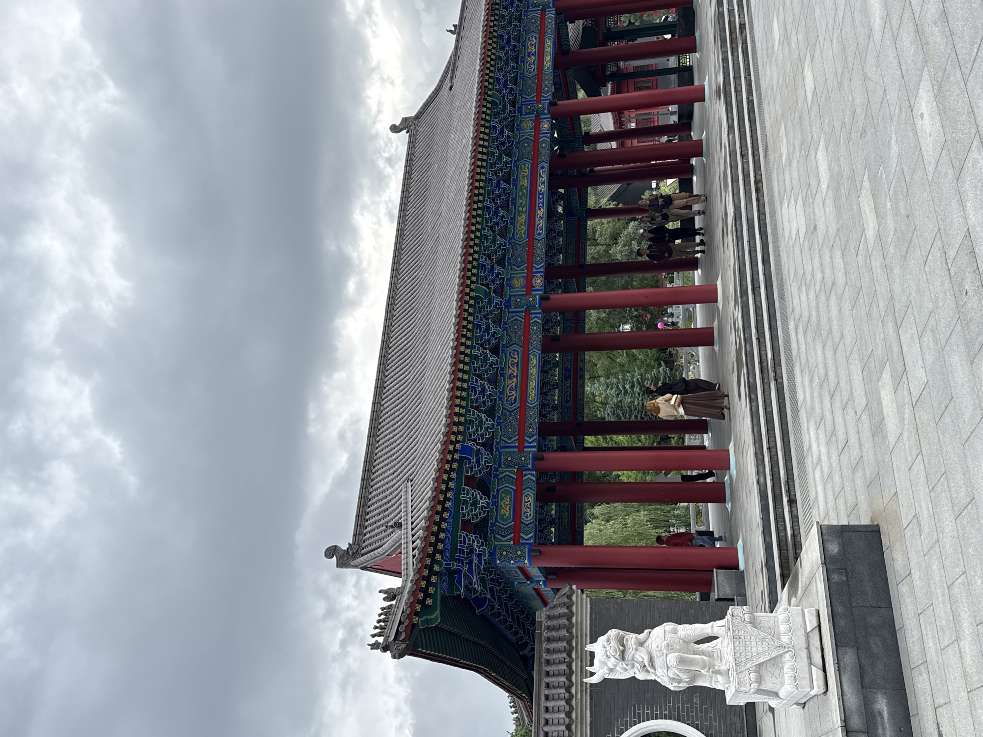 A wide, eye-level shot captures a large, traditional Chinese-style pavilion with a grey tiled roof, red cylindrical columns, and intricately painted eaves in vibrant blues, greens, and reds. The pavilion is set against a backdrop of overcast grey skies and lush green trees. In the foreground, to the left, stands a white stone Foo Dog sculpture on a tiered pedestal. Several people are visible beneath and near the pavilion, some standing and others walking, appearing to be tourists or visitors. The ground is paved with large grey square tiles. A wall made of grey bricks with a circular opening is visible to the left of the Foo Dog. The scene appears to be in a park or garden setting. This image was taken in the Chinese Garden of Friendship in Moscow, Russia, known for its traditional Chinese architecture and sculptures.