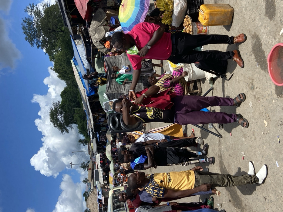 The scene is a bustling market area in Dar es Salaam, Tanzania, located at the coordinates -6.787 latitude and 39.113 longitude. A group of men and women are gathered around a large speaker, possibly selling merchandise or engaging in a performance. The man in the pink shirt is standing in front of the speaker, addressing the crowd. He is holding a microphone, and several people are looking at him attentively. The man wearing a purple shirt and a basketball jersey is holding a red cloth in his right hand. Several other individuals are standing nearby, observing the activity. Behind the group, there are stalls and vendors selling various goods. The market stalls have a variety of colorful fabrics, clothing, and goods. The overall atmosphere of the scene is bustling and vibrant, a typical sight in a Tanzanian market.