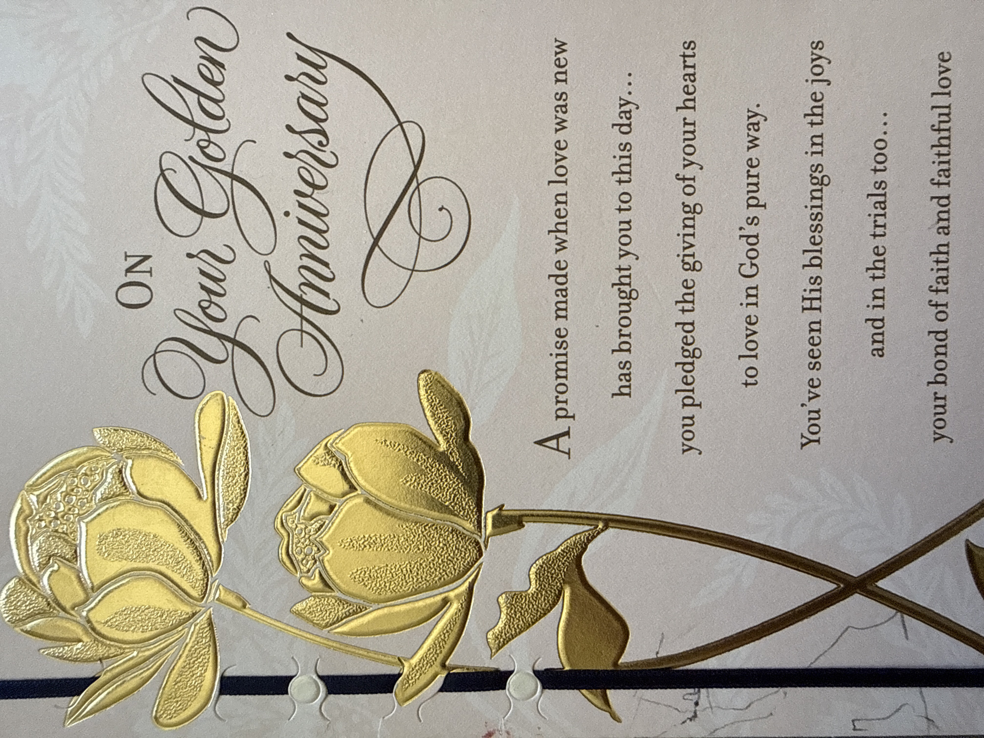 This is a close-up, eye-level, studio photograph of the front of a golden anniversary greeting card. The card features embossed gold floral designs and script text. The top portion of the card has a large, embossed gold flower with textured petals. Below it, a smaller embossed gold flower is visible. To the left of the flowers, a dark blue ribbon with light gray decorative circles is attached. The text on the card reads: "ON Your Golden Anniversary A promise made when love was new has brought you to this day... you pledged the giving of your hearts to love in God's pure way. You've seen His blessings in the joys and in the trials too... your bond of faith and faithful love". There are no people, actions, or interactions depicted. The environment is a studio setting for a product photograph. There is no information about the location of West Sacramento, United States of America.