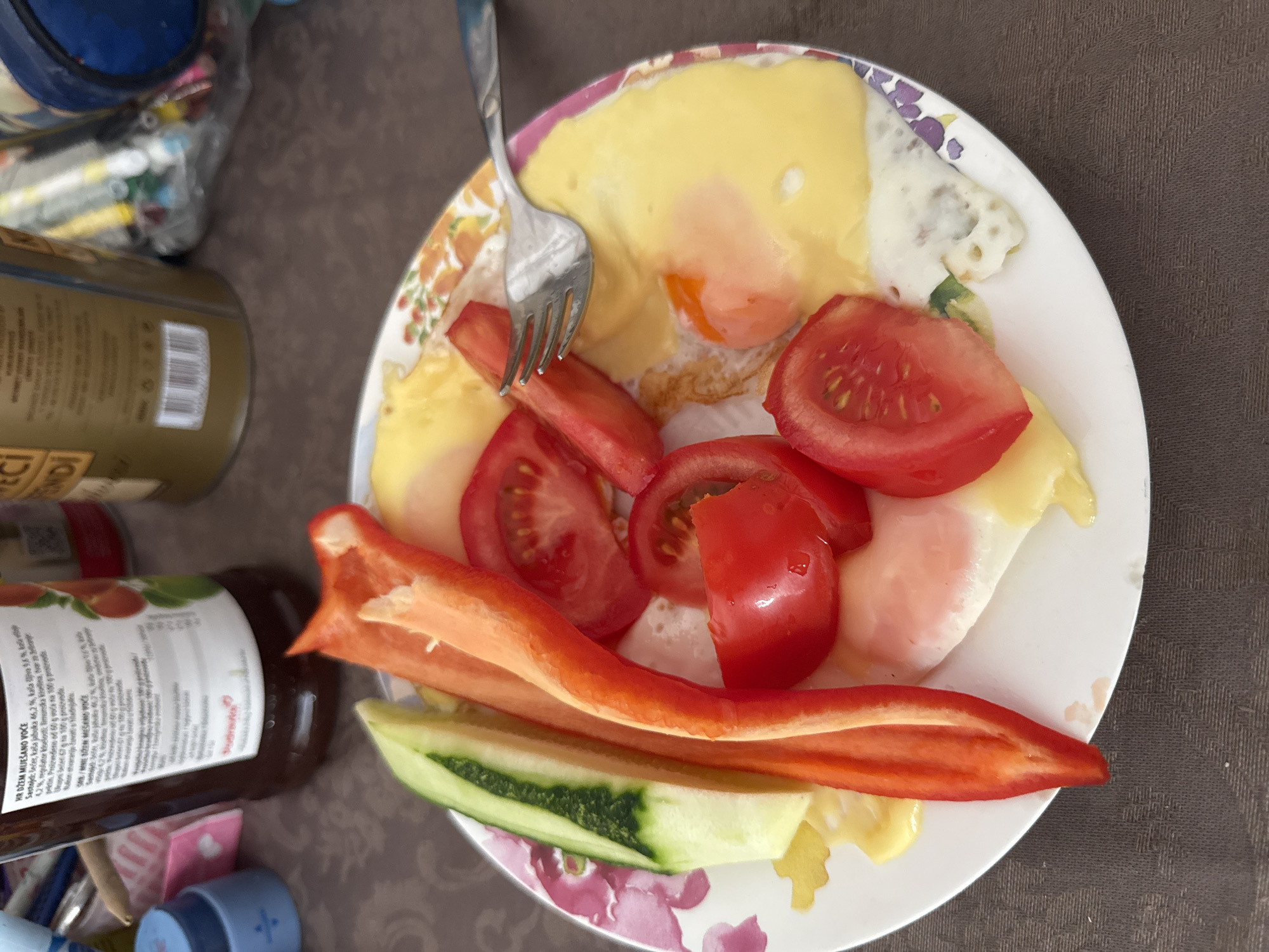 In Budva, Montenegro, a plate of food sits on a brown tablecloth. The plate contains fried eggs, sliced tomatoes, cucumber sticks, and red bell pepper strips. A fork rests on the plate, partially covering one of the fried eggs. In the background, several bottles and containers are visible, including a bottle with a label featuring fruit. The scene appears to be an indoor setting, possibly a dining area.