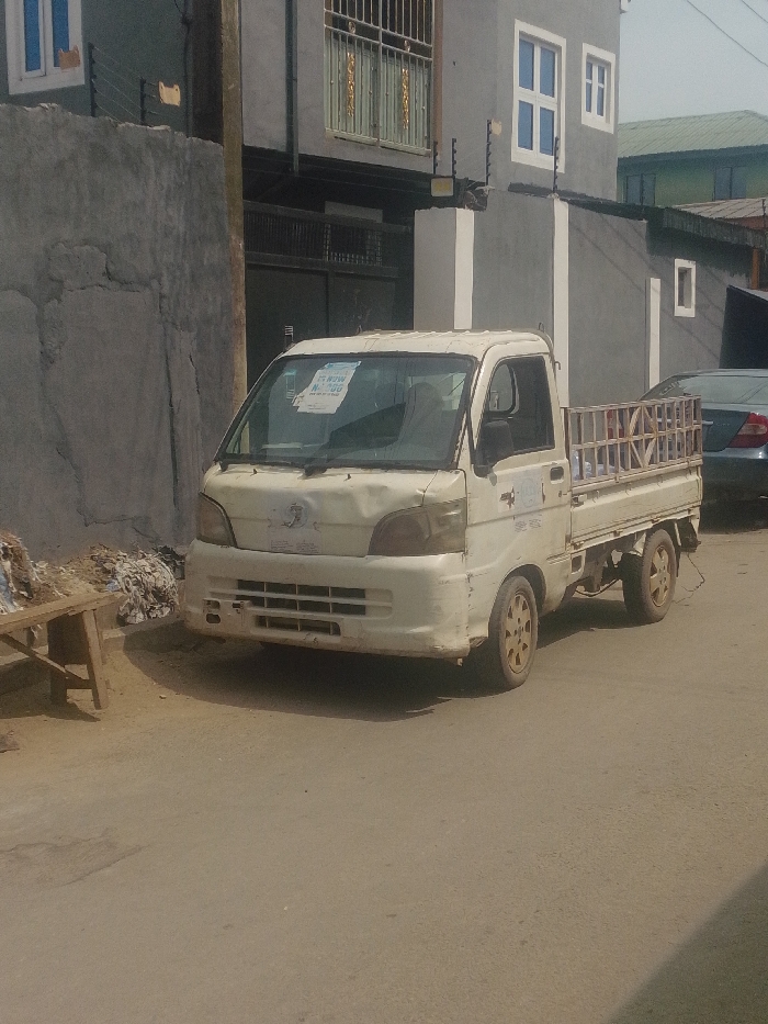 This image depicts a street scene in Agani, Nigeria, under daylight conditions. The primary subject is a white, four-wheeled, single-cab mini-truck with a flatbed, positioned on an asphalt road in the foreground. The truck exhibits signs of wear, including dents on its front hood, discoloration, and dirt accumulation on its wheels and body. A light blue and white "FOR SALE" sign, partially obscured but showing "N 700,000", is affixed to the inside of the front windshield.
To the immediate left of the mini-truck, a rudimentary wooden table is visible, upon which various discarded items, possibly fabric scraps or refuse, are piled. Behind the table stands a rough-textured concrete wall. A dark utility pole is situated between this wall and the background buildings.
In the background, a multi-story building with a grey facade and white-framed windows is prominent. This building features security fencing, appearing to be electric, mounted atop its perimeter wall. Another darker-colored building with barred windows is partially visible further back and to the left. To the right of the scene, behind the mini-truck, the front section of a dark-colored sedan is partially visible, parked facing away from the viewer. No individuals are present or discernible in the frame.