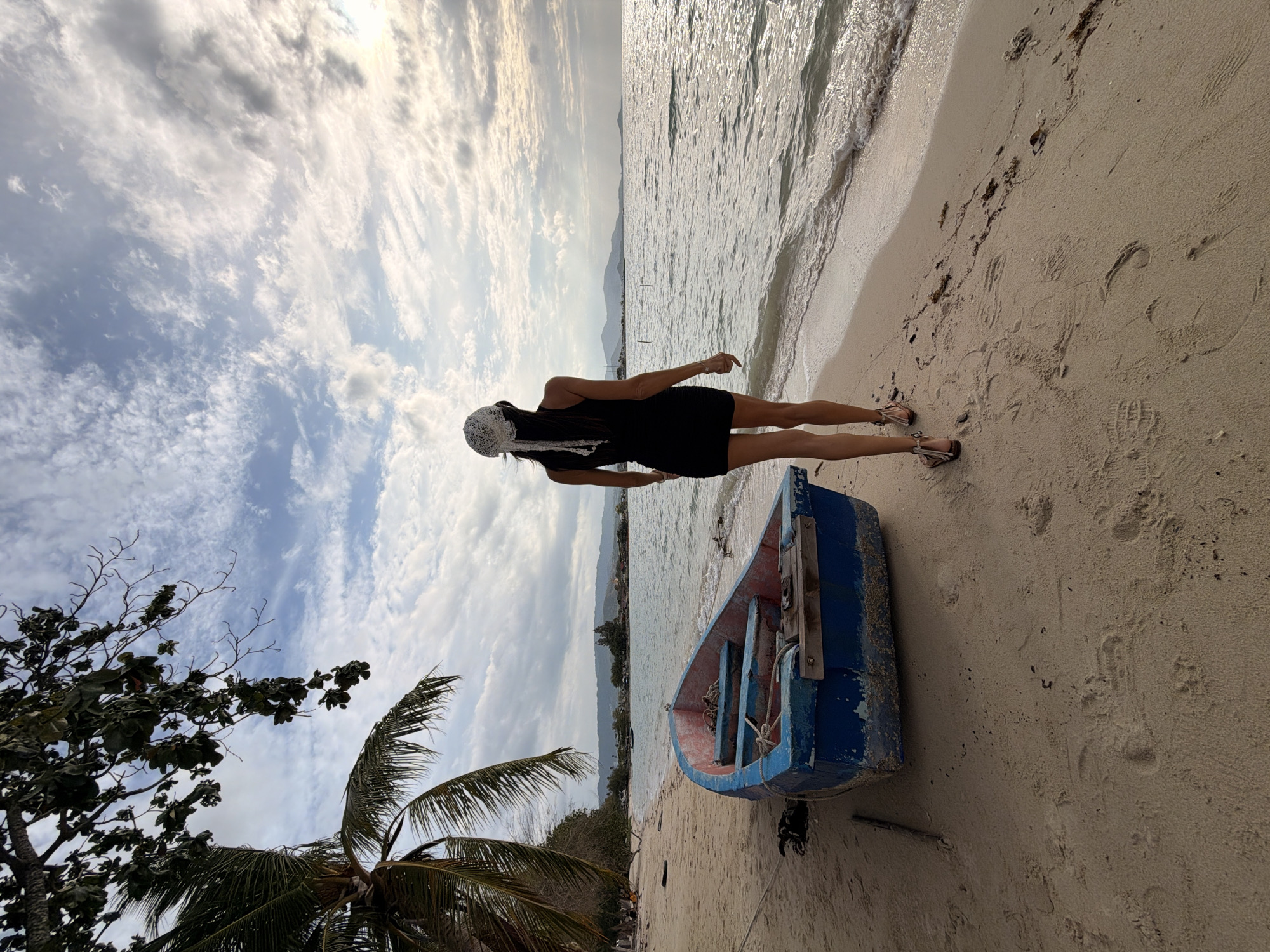 A woman in a black dress and a white headscarf walks on a sandy beach towards the ocean on the island of Ko Samui, Thailand. She is facing away from the camera, with her back to the viewer. The sun is setting, casting a warm glow on the scene. A blue and red rowboat sits on the sand to her left. Palm trees are visible in the background. The water is calm, with small waves lapping at the shore. The sky is filled with dramatic clouds.