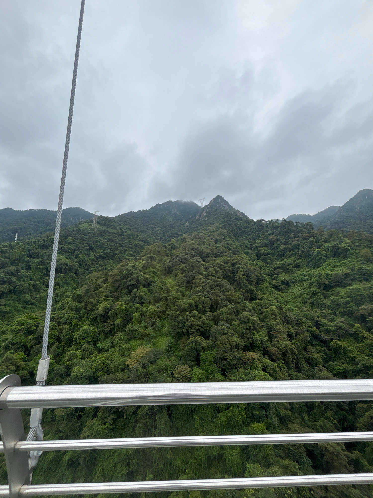 The image shows a lush, mountainous landscape covered in dense green foliage. The sky is overcast with heavy gray clouds. In the foreground, a metallic railing with multiple horizontal bars is visible, suggesting a viewing platform or structure. A thick metal cable runs vertically along the left side of the frame, secured by metal fixtures to the railing. The mountains rise steeply, dotted with what appear to be power line towers in the distance. The scene conveys a sense of elevation and natural scenery. This scene is likely from the Xiqiao Mountain area in the Nanhai District of Foshan, China, known for its scenic beauty and cable car service which would explain the presence of the railing and cable. There are no people visible in the image.