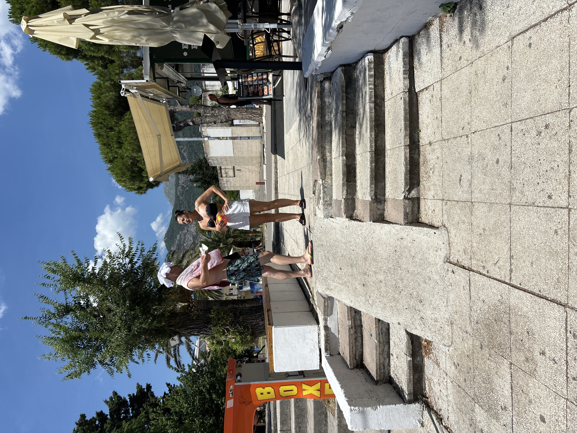 Two individuals are standing on a concrete ramp and steps in Budva, Montenegro, on a sunny day. The man, on the left, wears patterned swim trunks and a white towel wrapped around his head and shoulders. He is holding a small, white object in his hands and appears to be talking to the woman. The woman, on the right, is wearing a black bikini top and white shorts. She is holding a colorful beach ball and looking at the man. Behind them, to the left, is an orange structure with yellow letters spelling "BOX." To the right, there is a cafe with outdoor seating and umbrellas. In the background, there are trees and buildings, with a mountain range visible in the distance. The scene suggests a casual, outdoor setting likely near a beach or tourist area.