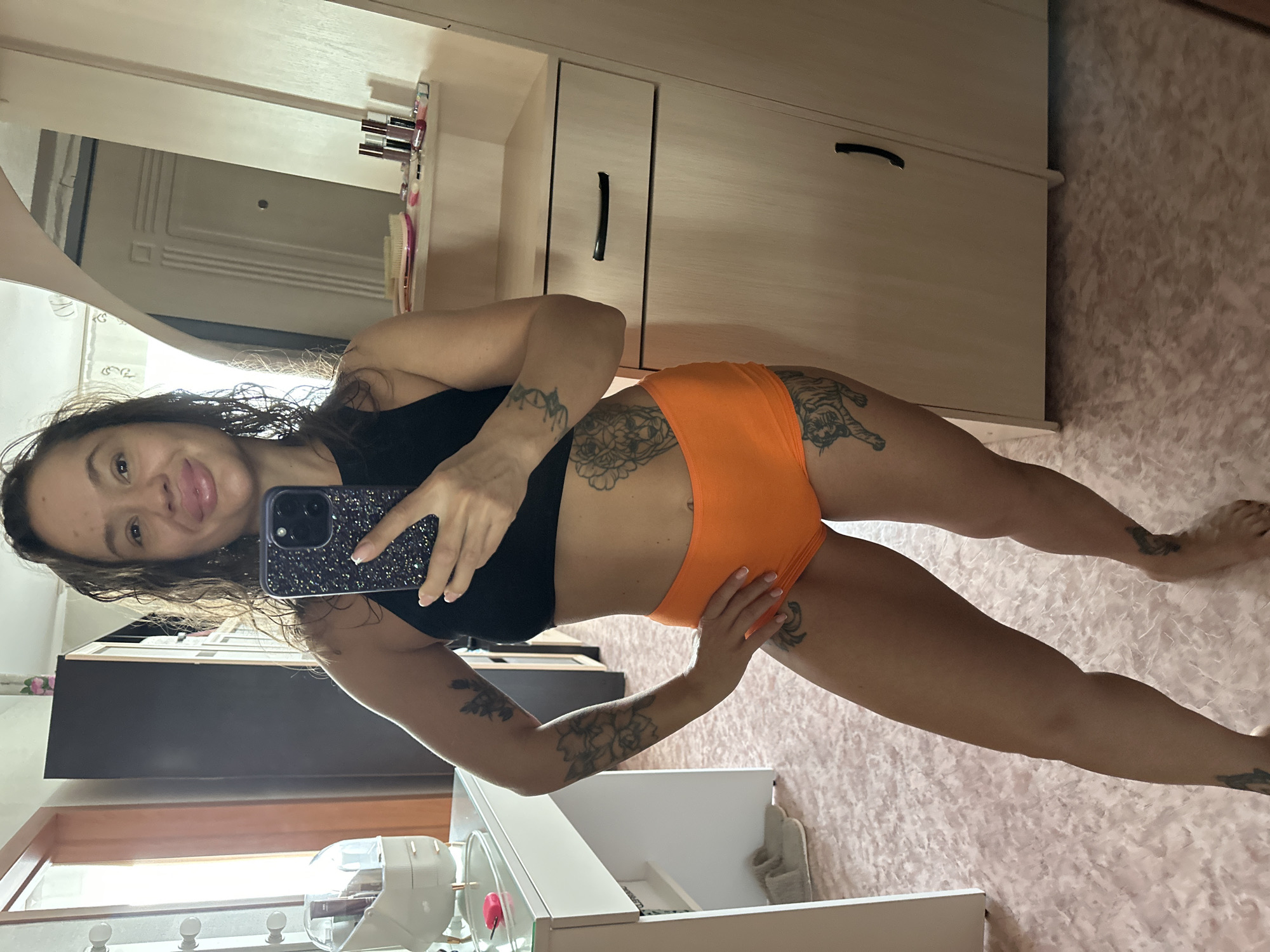 A woman is taking a mirror selfie in a room. She has dark curly hair and is wearing a black crop top and orange shorts. She has tattoos on her arms, torso, and legs. The room appears to be a bedroom or dressing area, with a vanity table on the left and a closet or wardrobe on the right. A mirror reflects the woman and part of the room. The floor has a pink patterned carpet. The overall impression is that of a casual, private setting. There is no indication of the city of Yuzhnouralsk, Russia.