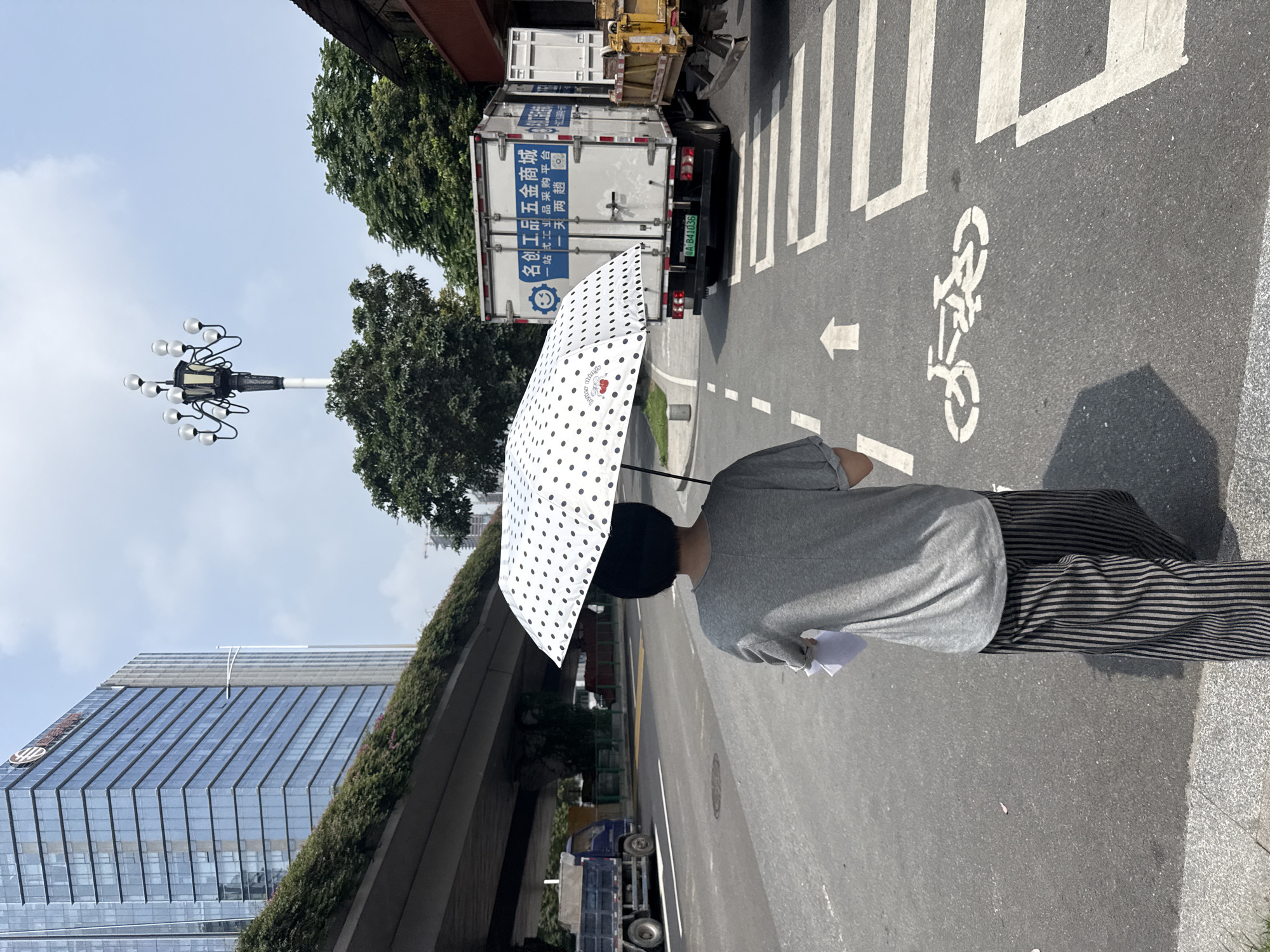 A person walks away from the camera on a city street, holding a white polka-dotted umbrella over their head. They are wearing a grey t-shirt and black and grey striped pants. The person is walking on the sidewalk next to a road with traffic. In the background, there is a large modern building, a street lamp with multiple bulbs, trees, and a white truck with blue lettering. A crosswalk and a bicycle lane marking are visible on the road. The image appears to have been taken in Tianhe District, China, under a bright, cloudy sky.