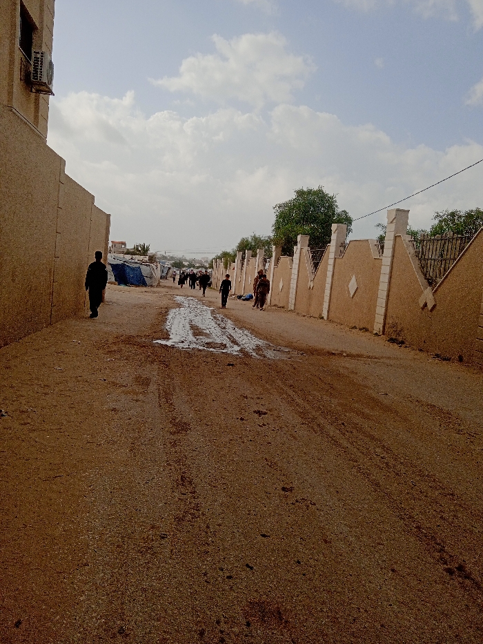 The image depicts a dirt road in Khan Yunis, Palestinian Territory, under a partly cloudy sky. The road surface is unpaved, showing tire tracks and a prominent muddy patch near the center of the frame. On the left, a light tan building wall with a textured finish extends into the distance, featuring an external air conditioning unit mounted high on its initial section. On the right, a decorative light tan wall with repetitive white or light-colored pillars and diamond-shaped inserts runs parallel to the road, topped by sections of metal fencing and partially obscured by green foliage. Numerous individuals, estimated at 8-10, are observed walking away from the camera along the road. One person in dark clothing is on the far left, near the building wall. Further down the road, a group of people, some appearing to carry bundles, are visible walking towards structures constructed from blue tarpaulins and other materials, suggesting temporary shelters or a camp in the middle distance.
