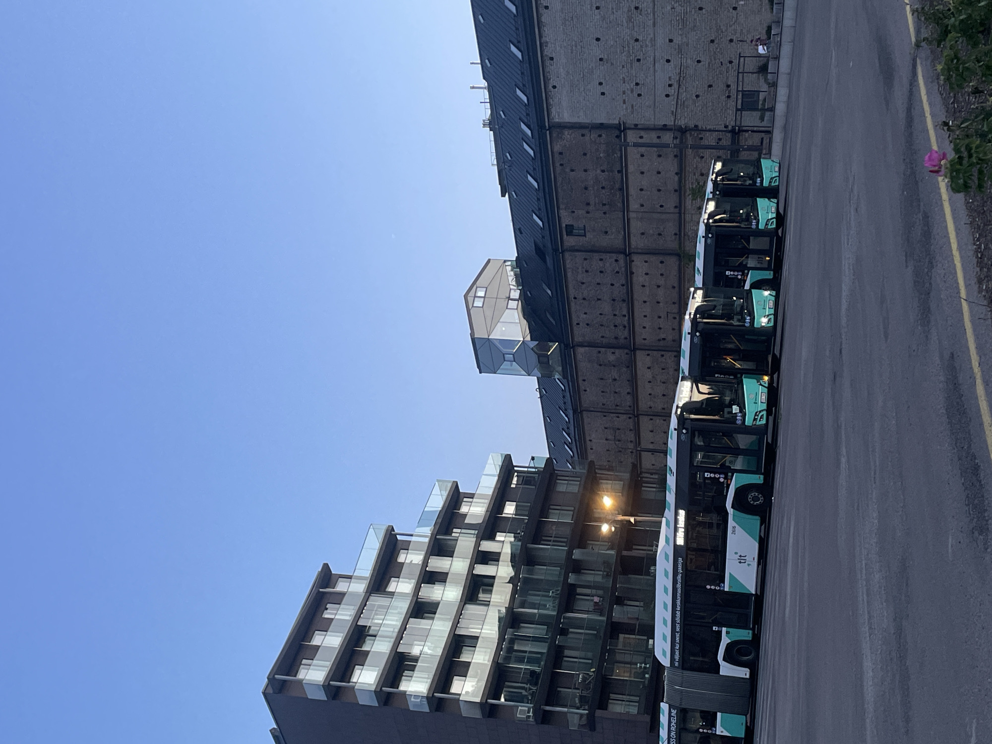 The image depicts a clear sky over a cityscape in Tallinn, Estonia. In the foreground, several modern buses are parked in a lot. The buses are predominantly white with teal accents and branding. To the left, a tall, multi-story apartment building with glass balconies stands out. Behind the buses and to the right, a long, low brick building with a sloped, dark roof punctuated by dormer windows is visible. A unique, angular glass structure is situated atop this brick building. The overall scene suggests an urban environment, possibly a bus depot or a street in a developed area of the city. Two individuals are faintly visible in the far right background, appearing to be standing near the brick building.