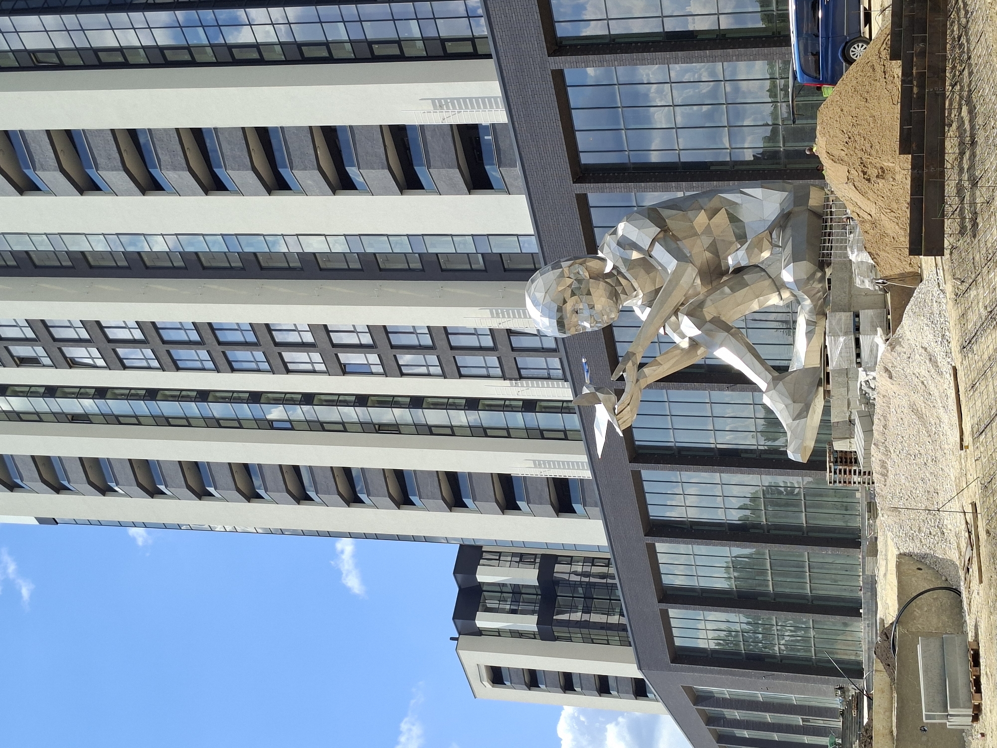 The image depicts a modern, geometric metal sculpture of a child holding a toy airplane, seated in front of a newly constructed, multi-story building with a dark brick base and light-colored facade. The building features numerous glass windows and balconies. In the background, another building of similar architectural style is visible. The foreground shows a construction site with piles of sand, gravel, concrete blocks, and rebar, indicating ongoing development. A blue van is parked to the right of the sculpture. The sky is blue with scattered clouds.
The sculpture and building are located in the "Alley of Moldovan Classics" in Chișinău, Moldova.