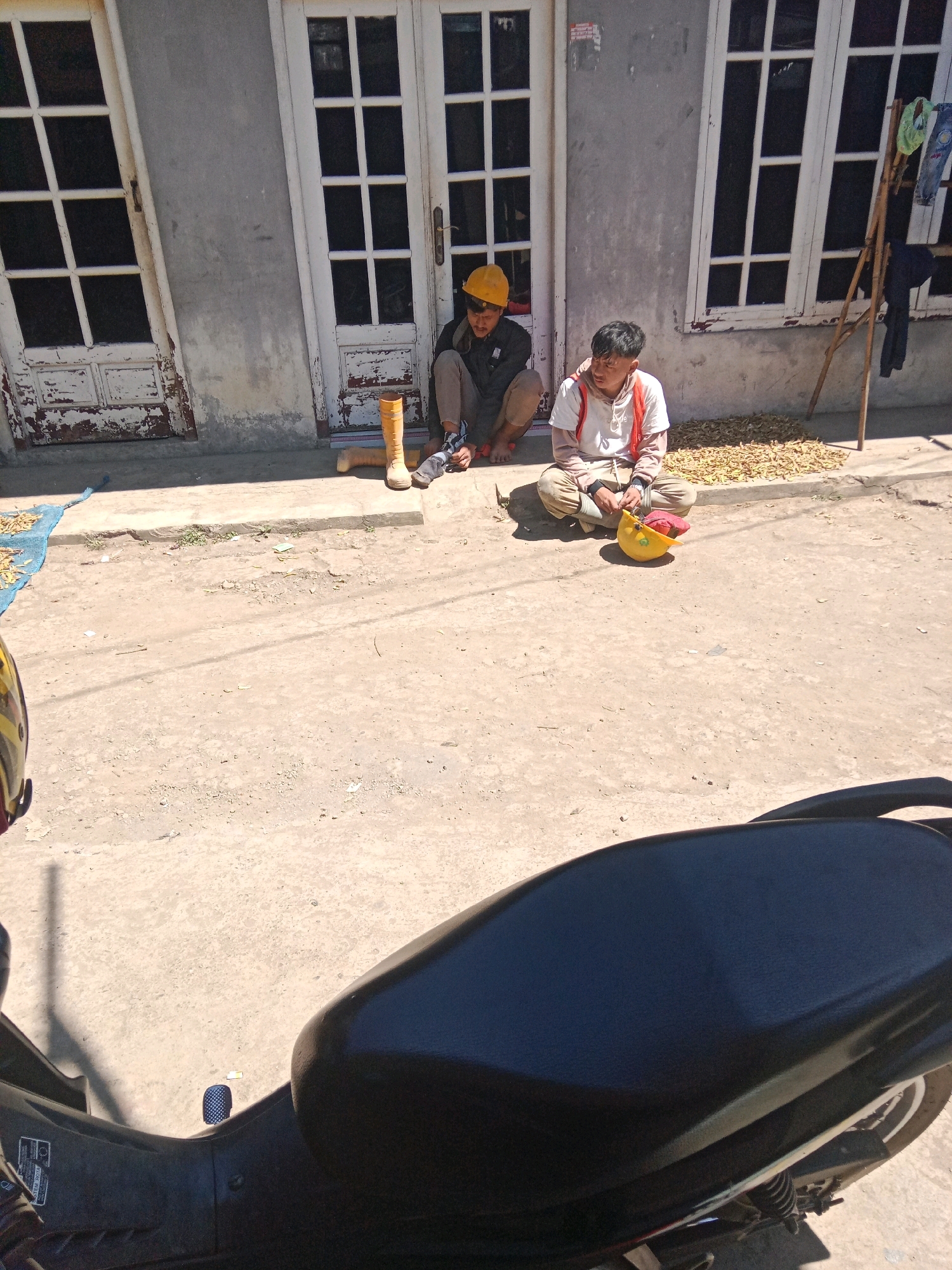In Jurangjeru, Indonesia, two young males are seated on the ground in front of a building with distressed white double doors. The male on the left wears a yellow hard hat, a dark jacket, and tan pants. His left leg is bent, with his foot resting on an orange boot. His right leg is also bent, and he appears to be tying his shoe. To his right, the second male sits with his legs crossed. He wears a white t-shirt with an orange vest and light-colored pants. In front of him is a yellow hard hat and a red object, possibly a bag or hat. Behind the seated individuals, a pile of what appears to be dried beans or pods is spread on the ground, with a blue tarp partially visible to the left. A bamboo drying rack with clothes hanging on it is situated to the right of the building. The ground is dusty and appears to be a dirt road or pathway. In the foreground, the black seat of a motorcycle is prominently visible, obscuring the lower portion of the scene.