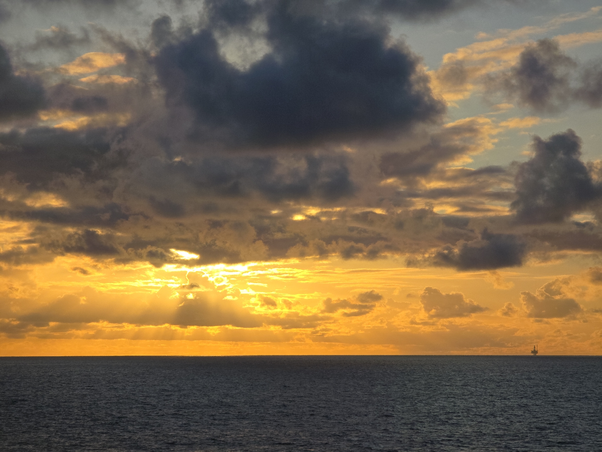 The image shows a sunset over the ocean. The sky is filled with dark and golden clouds, with the sun shining through in the center of the horizon. The ocean is dark and calm. In the distance, a small oil rig is visible. The location is unknown.