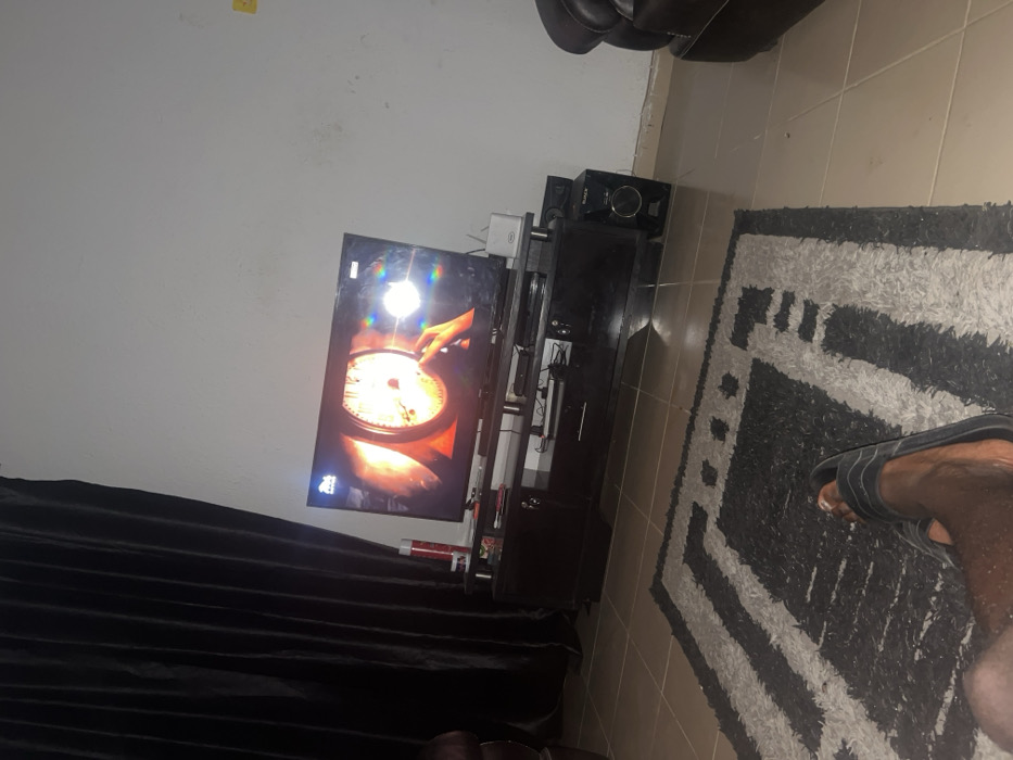 A living room features a television mounted on a black entertainment center with a speaker atop. The TV displays a close-up of a cooking scene. Dark curtains partially obscure the bottom left of the image. A rug with a black and white pattern is positioned on the tiled floor. A foot in a sandal is visible on the rug's edge. The scene is captured from an angle, giving a slightly tilted perspective.