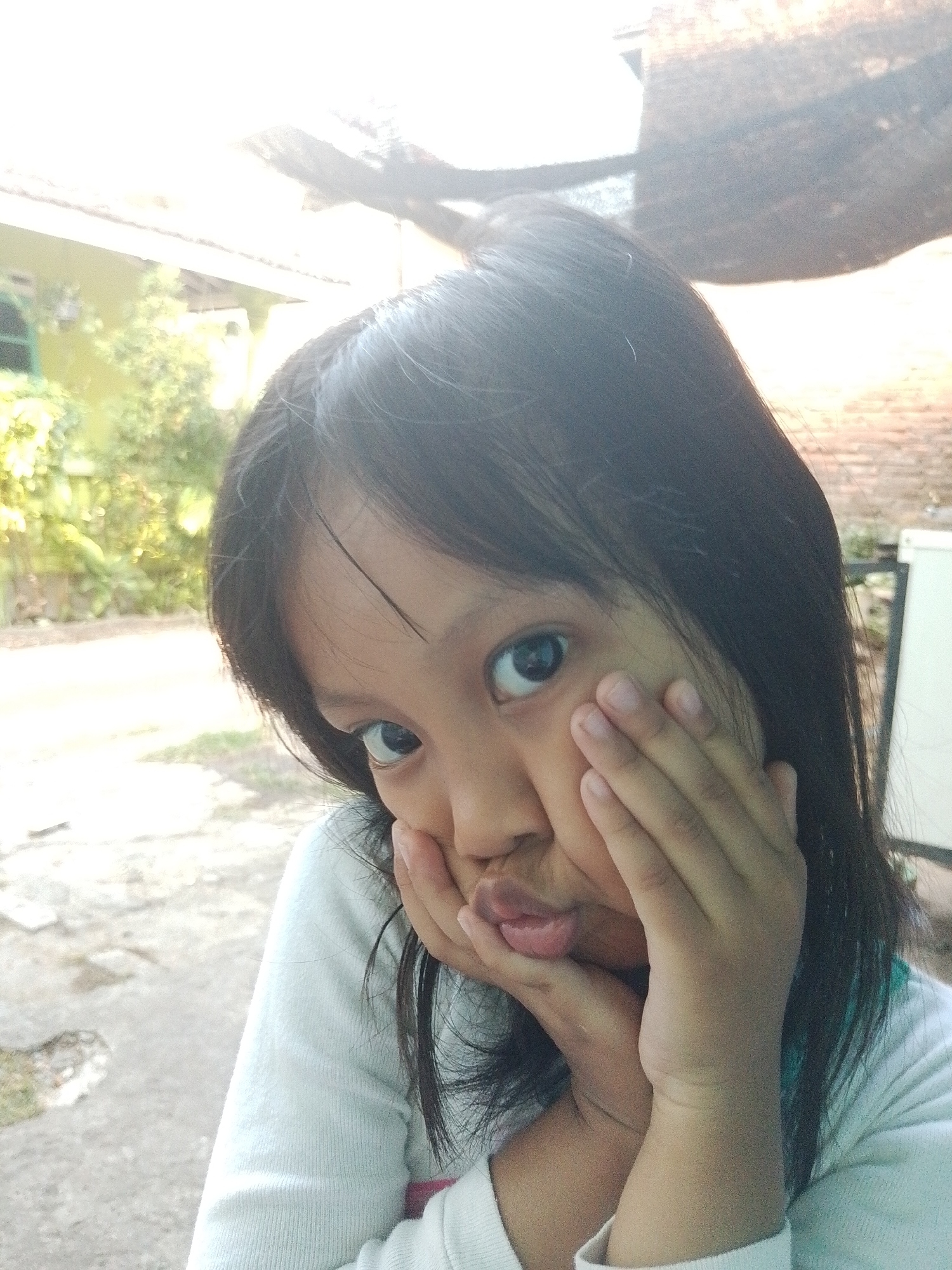 A young girl with dark hair pokes her tongue out and pushes her cheeks in with her hands, making a funny face. She is wearing a light blue long-sleeved shirt. The background appears to be an outdoor setting with a house and trees visible, possibly in a residential area of Kecamatan Pati, Indonesia. The lighting suggests it is daytime, with sunlight creating some glare in the upper portion of the image.