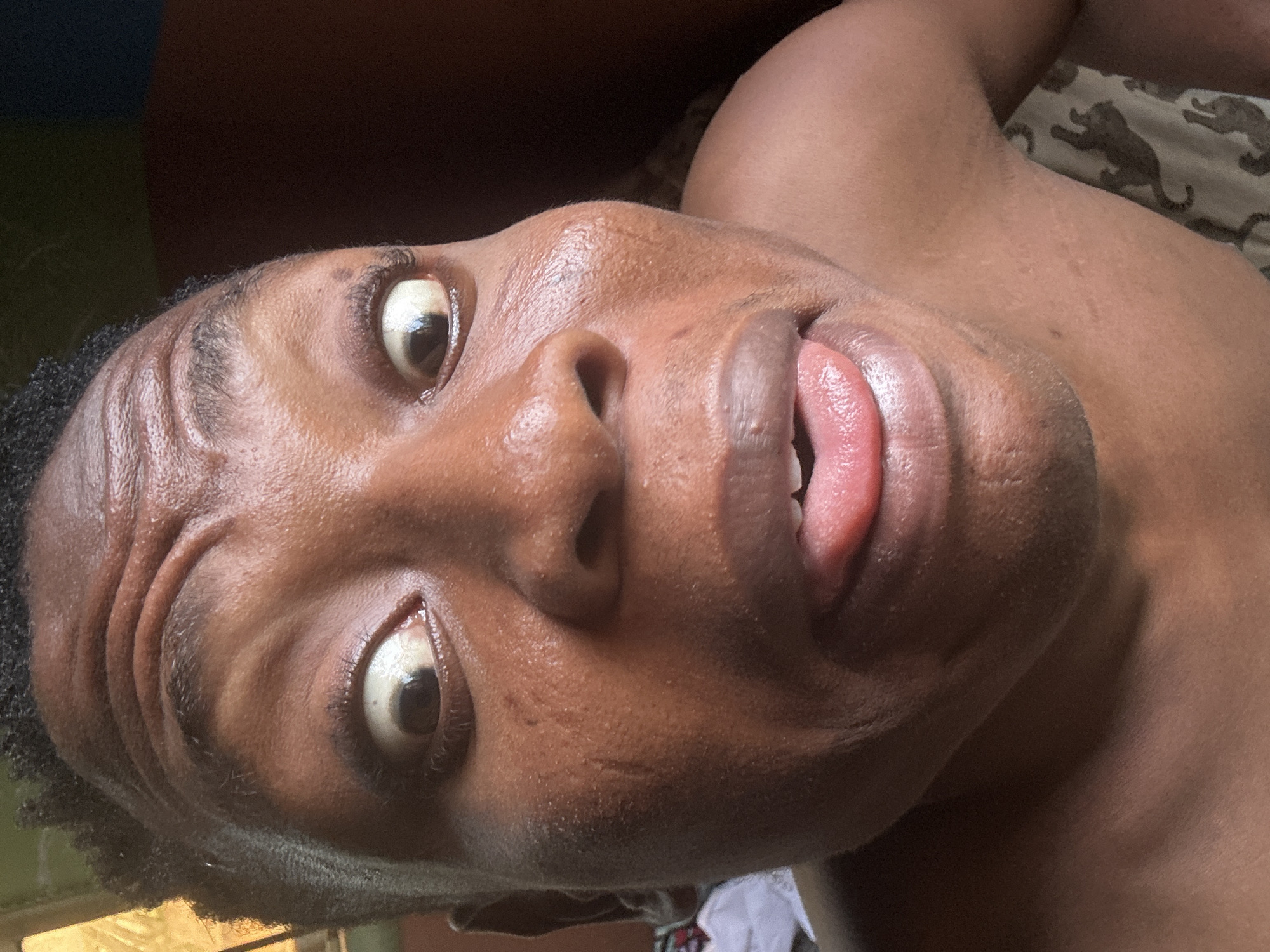 The image is a close-up selfie of a Black individual with their tongue sticking out. The person's eyes are wide open, and their forehead is wrinkled. The skin appears to have some texture and a slight sheen, possibly due to sweat. The background is dark and out of focus, with hints of a brown surface on the right and a lighter, possibly patterned fabric on the lower right. The image is taken in Ikorodu, Nigeria.