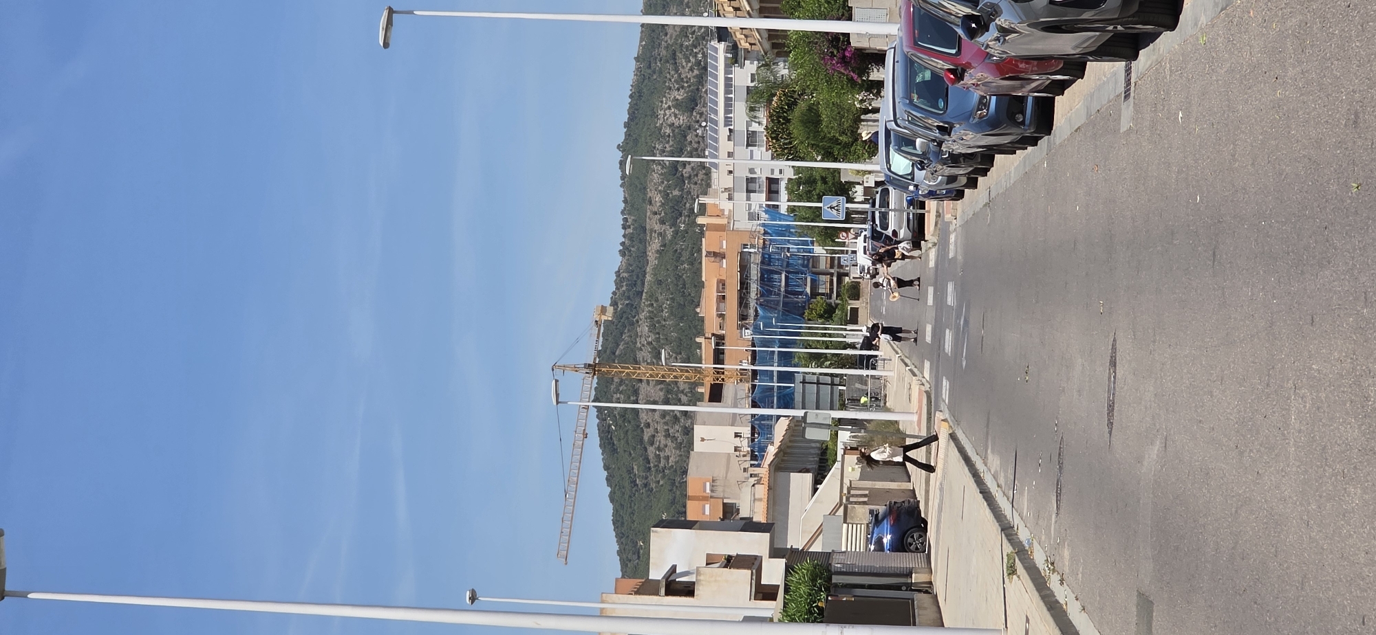 The image depicts a street in Sagunto, Spain, lined with parked cars on the right side. A construction crane is visible in the background, behind a row of buildings. Several people are walking in the street, and one person is walking on the sidewalk. Streetlights line the left side of the road, extending towards the sky. Hills covered with vegetation are in the distance. The sky is clear and blue.
