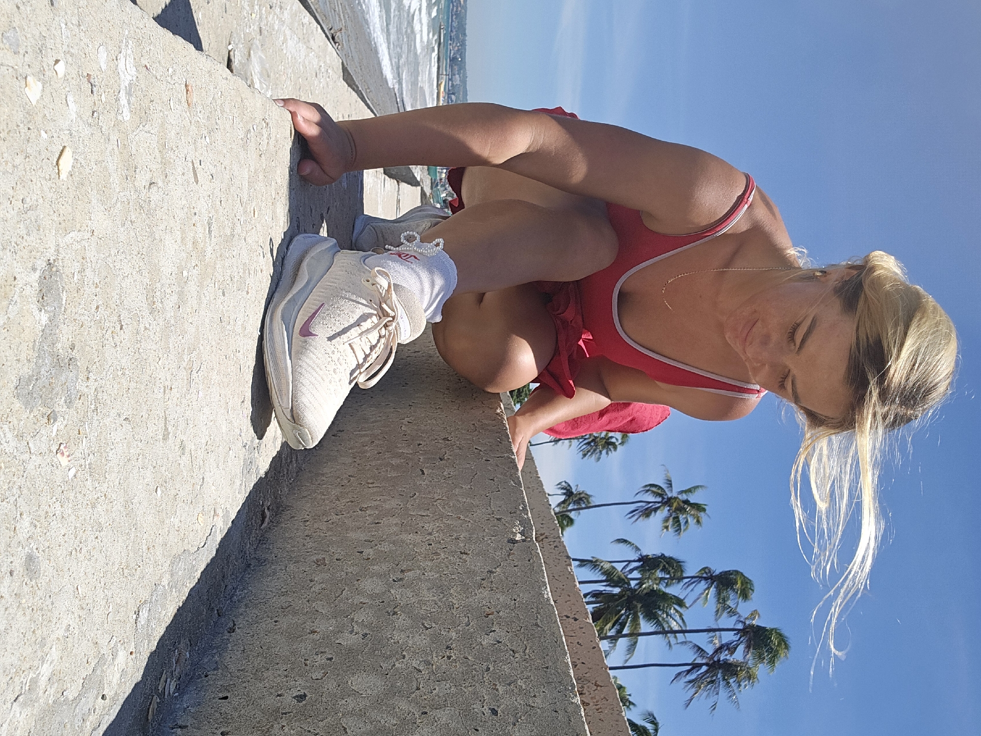 A young blonde woman in a red swimsuit is crouching on a concrete embankment in Mui Ne, Vietnam. She is looking down at her right foot, which is clad in a beige athletic shoe and white socks. Her hair is blowing in the wind. In the background, the ocean and palm trees are visible under a clear blue sky. The embankment appears to be part of a coastal walkway or sea defense. The lighting suggests it is daytime, likely mid-morning or afternoon given the bright sunlight and shadows.