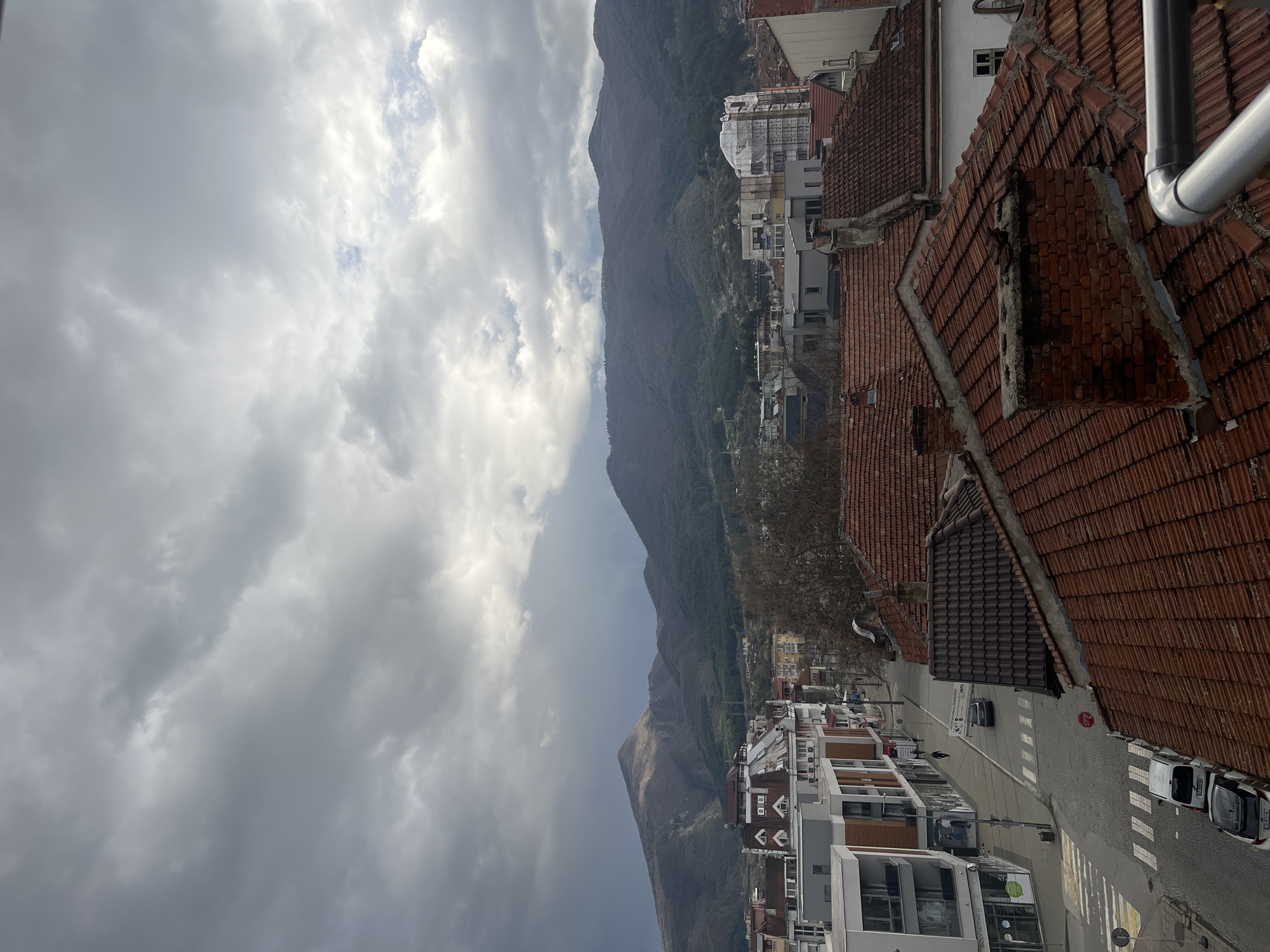 The image depicts a view of the city of Goce Delchev, Bulgaria, under a cloudy sky with mountains in the background. The foreground consists of red-tiled rooftops of buildings, and a street with a pedestrian crossing, cars, and buildings on either side. The street runs perpendicular to the mountain range, showcasing a blend of urban and natural elements.