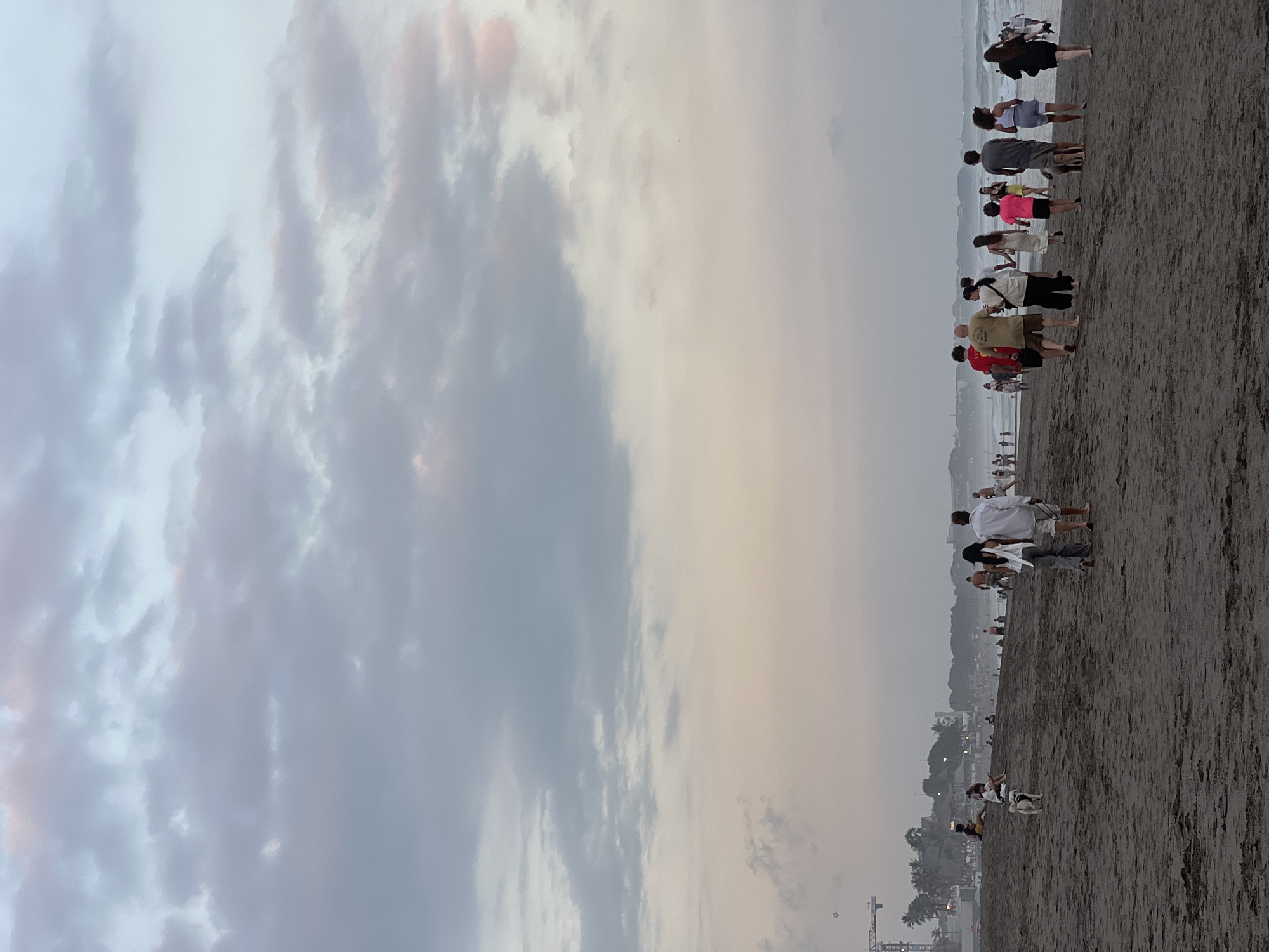 This image depicts a beach scene in North Kuta, Indonesia, during what appears to be either sunrise or sunset. The sky is filled with a mix of gray, white, and pale pink clouds, with a lighter pastel hue near the horizon. The foreground is dominated by dark, wet sand, showing footprints from people walking.
In the mid-ground, a group of people are walking along the shoreline, most facing away from the camera and towards the ocean. They are dressed in casual beachwear. Some individuals are walking alone or in pairs, while others are in larger groups. One person appears to be walking a dog. Further in the background, the ocean is visible with gentle waves and some people wading in the water.
Along the distant coastline, a line of palm trees and buildings is visible, suggesting a developed area. A construction crane is also present on the left side of the frame, indicating ongoing development. The overall atmosphere is serene and relaxed, characteristic of a beach at dawn or dusk.